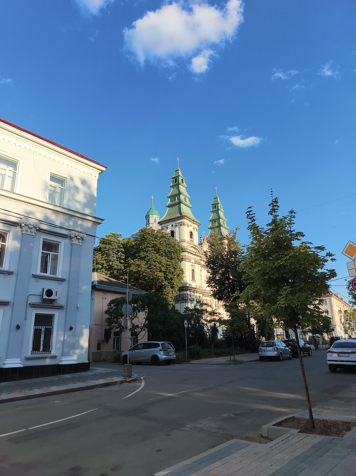 The image shows a street scene in Ternopil, Ukraine. In the foreground, a light blue building with white trim is on the left, and a street runs diagonally across the image. Several cars are parked on the right side of the street. In the background, a large white church with green spires is visible behind a cluster of trees. The sky is blue with some white clouds.
