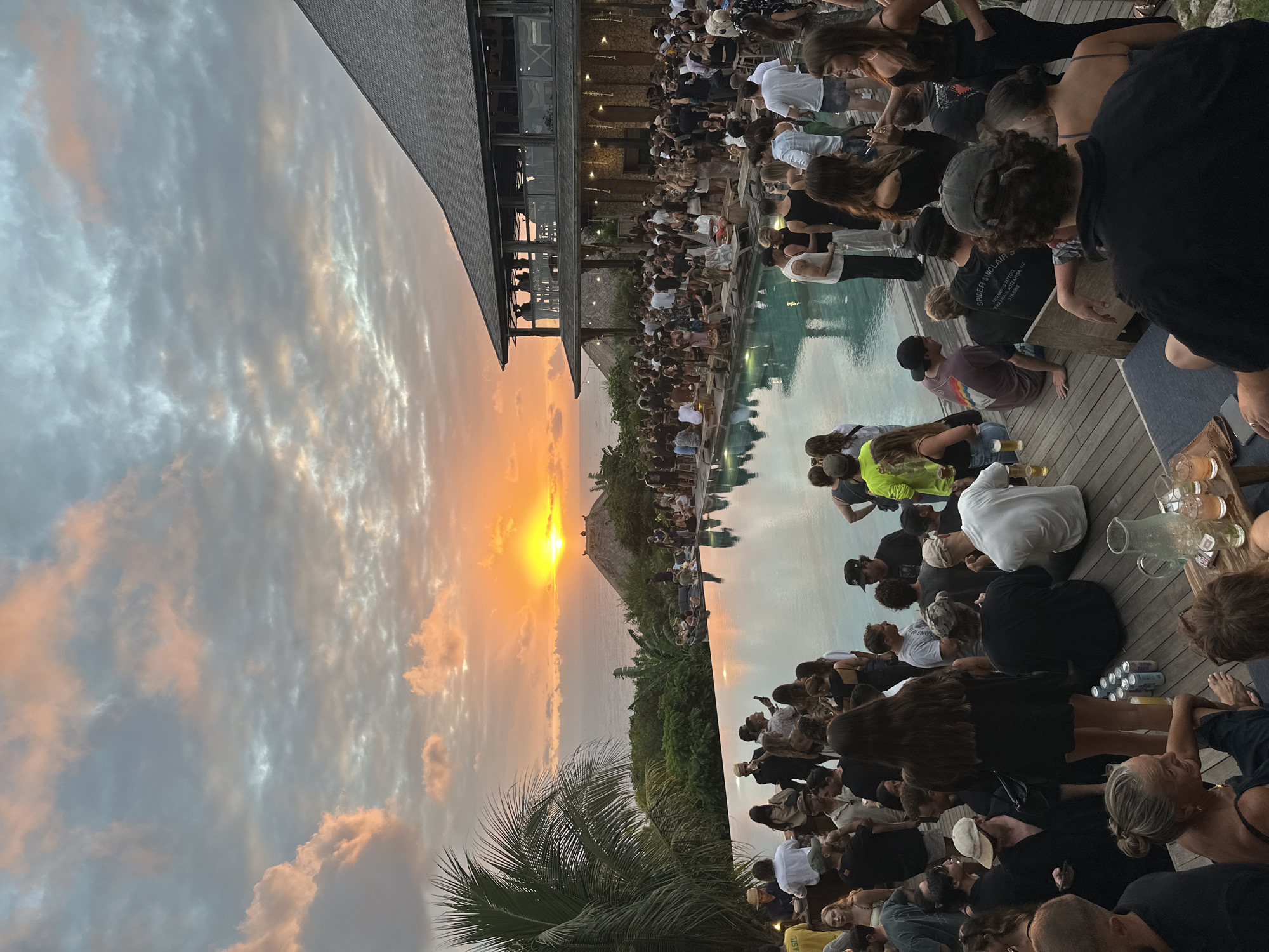 In Kuta Selatan, Indonesia, a large group of people are gathered for an event at a resort during sunset. The setting sun casts a warm glow over the scene, illuminating a cloudy sky with hues of orange, pink, and gray. A large infinity pool dominates the foreground, reflecting the vibrant sky and the surrounding lush greenery. Several individuals are seated around the pool on wooden loungers and benches, while others stand and mingle.
A multi-story building with a pitched roof, likely part of the resort, is situated to the right of the pool, featuring balconies and open-air spaces where more people are visible. String lights hang from the building, adding to the festive atmosphere. The architecture appears to be made of natural materials like wood and stone.
The crowd is diverse, with people dressed in casual attire, suggesting a relaxed social gathering. Some are holding drinks, and cans of beverages are visible on a wooden table near the foreground. The overall impression is one of a lively party or social event taking place in a beautiful tropical setting, coinciding with the dramatic sunset over the ocean. The ocean is visible in the distance, beyond the resort and the coastline.