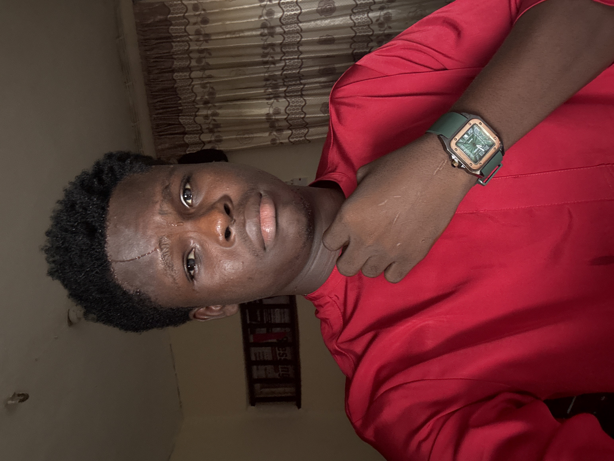 A young Black man with short, dark, curly hair is shown in a close-up portrait, looking directly at the camera. He has dark brown eyes and a light brown complexion, with a scar visible on his forehead. He is wearing a vibrant red long-sleeved shirt. His right hand is raised, with his fingers lightly touching his chin. On his left wrist, he wears a wristwatch with a square, rose gold-colored face and a dark green band.
The background consists of a dimly lit room. To the left, a framed object with what appears to be text and grid-like patterns hangs on a beige wall. To the right, patterned beige curtains with a floral design are drawn. The ceiling is visible at the top of the frame and appears to be painted a light color. The image suggests an indoor setting, potentially a home in Ihiagwa, Nigeria.