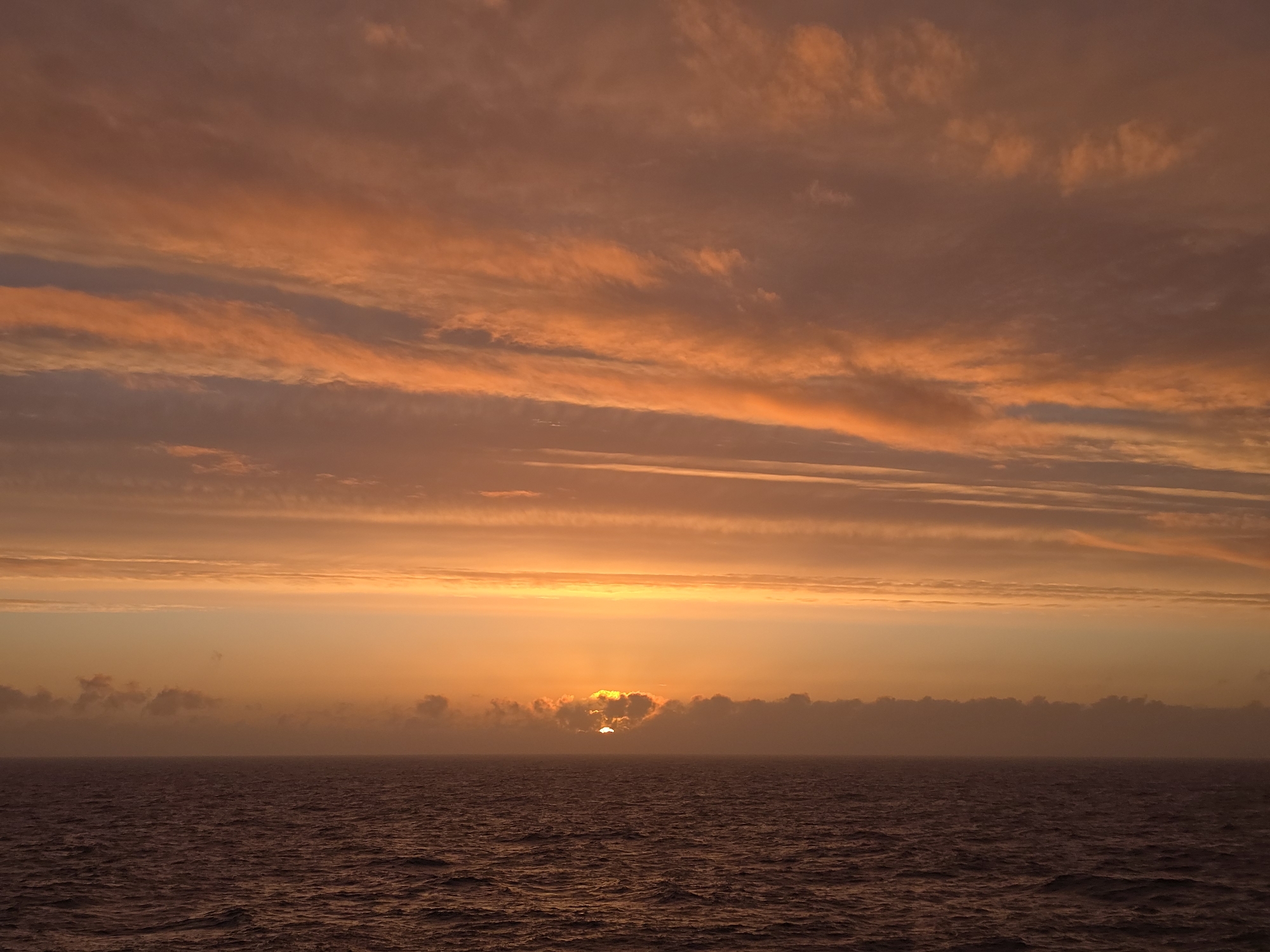 The image depicts a sunset over the ocean. The sky is filled with orange and red hues, with streaks of clouds. The sun is partially obscured by clouds near the horizon. The ocean is dark and calm, reflecting the colors of the sky. The location is Unknown, Unknown. There are no people visible in the image.