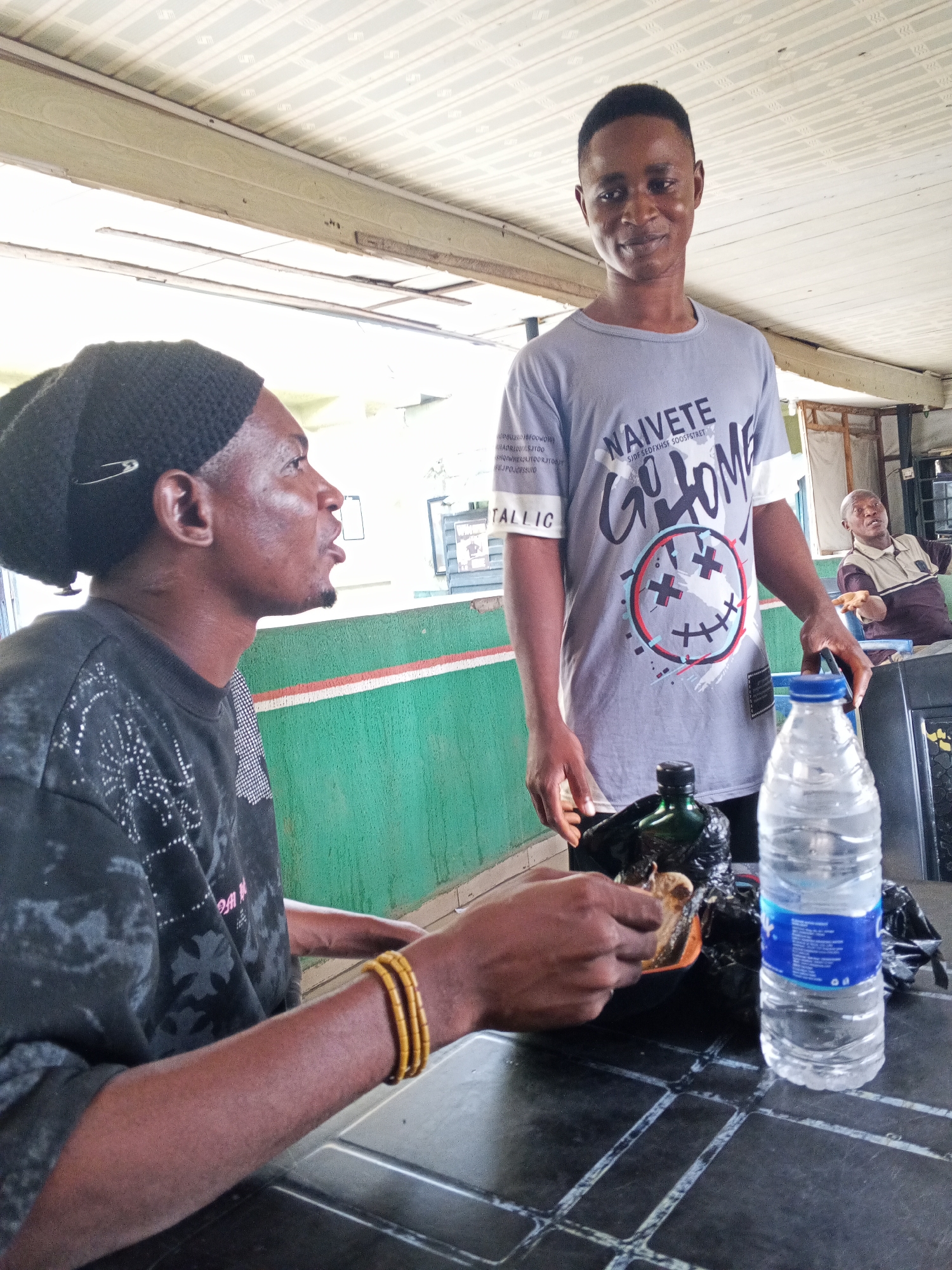 This image, taken in Zango-ndaji, Nigeria, portrays two men interacting in what appears to be a casual indoor or semi-open eating establishment during the daytime. A third man is visible in the background.
On the left, a man with dark skin is seated, captured in profile. He wears a black knitted beanie secured with a silver safety pin and a dark long-sleeved shirt with light patterns. His right wrist is adorned with multiple yellow-brown beaded bracelets. He is actively eating, holding a piece of food (possibly a pastry or bread) over an orange bowl, which rests within a black plastic bag. His mouth is slightly open, suggesting he is in the process of chewing or speaking.
Standing beside the table, facing the camera with a subtle smile, is another man with dark skin and short, dark hair. He wears a light gray t-shirt featuring prominent black text that reads "NAIVETE" and "GO HOME," accompanied by a large graphic of a stylized smiley face with 'X's for eyes. Smaller, less legible text includes "SJDF SEDFXHST SOOSFSTRET" and a partial "TALLIC" on his left sleeve. He is holding a black plastic bag with his right hand, resting on the table.
The dark, possibly plastic table has a subtle grid pattern. On it, between the two men, are a green bottle, a black plastic bag, and a clear plastic water bottle with a blue cap and a blue label.
In the background, a third man with light skin is seated at another table, looking to his right. The establishment features a green wall with a white and orange stripe, and a light-colored, patterned ceiling. The bright lighting suggests it is daytime. The overall atmosphere is relaxed and informal.