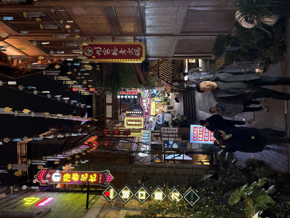 The image displays a brightly illuminated pedestrian alley at night, situated within Yuzhong District, China. Overhead, numerous decorative strings of wind chimes and lights are suspended across the passage. The right side of the alley features multi-story wooden buildings with traditional Chinese architectural elements, while the left side presents brick structures adorned with various neon signs. Prominent signage includes a large vertical red and yellow neon sign on the left with Chinese characters, and another similarly styled sign on the right advertising "Jiugongge Laohuo Hotpot." Additional illuminated signs for businesses, many displaying Chinese text and food imagery, are visible throughout. Potted plants and greenery are arranged along the base of the buildings on both sides. Approximately nine individuals are present, primarily engaged in walking along the paved alley, which slopes upwards towards a set of stairs in the background. In the immediate foreground, a male subject wearing a dark jacket and trousers stands, looking towards the viewer. Further into the alley, a large panda statue is visible near the base of the ascending stairs. The scene is densely lit by artificial sources from the commercial signs and overhead decorations, creating a vibrant nocturnal environment.