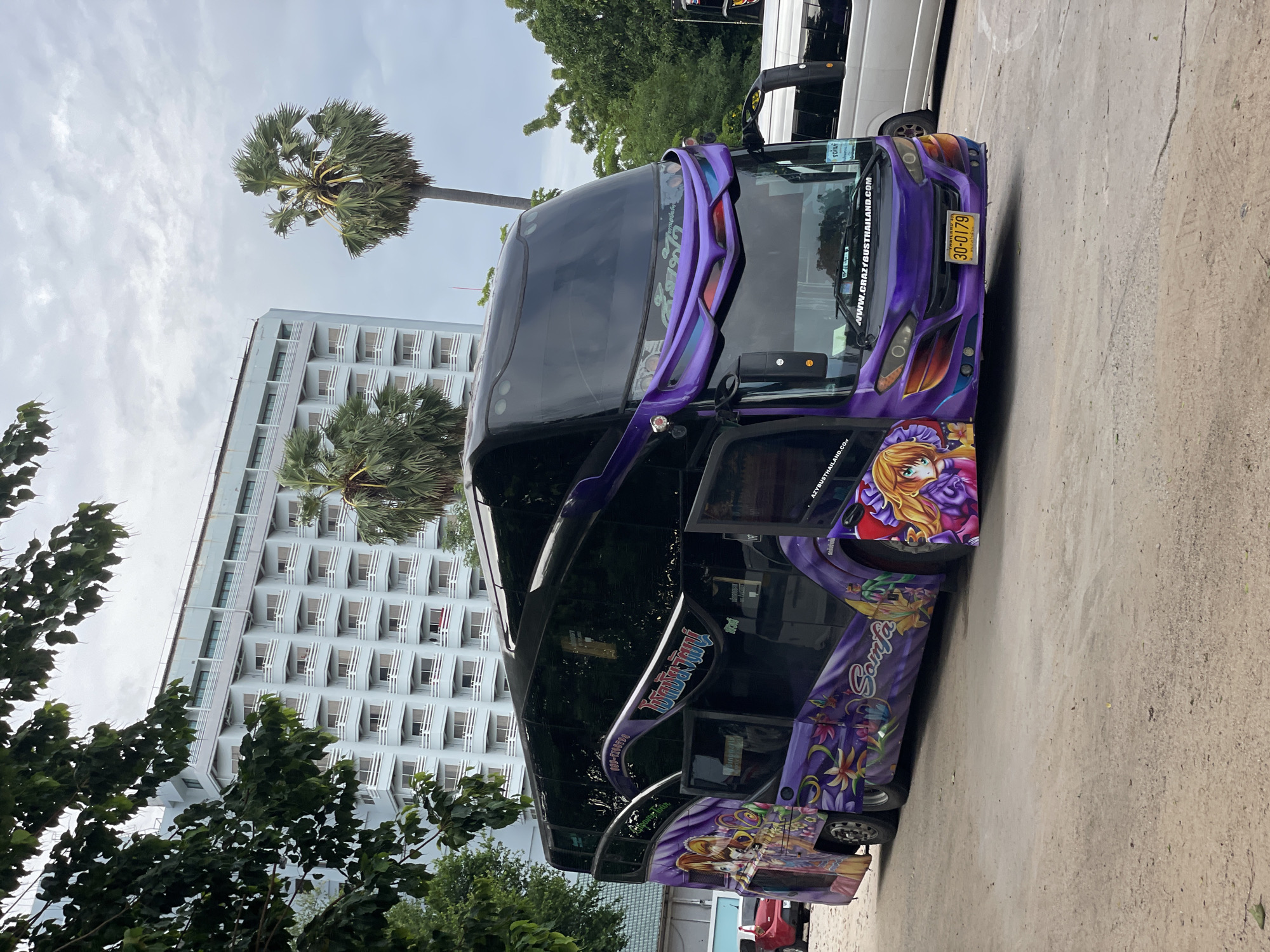 The image shows a purple double-decker bus parked in an outdoor area. The bus is decorated with anime-style characters and floral designs. The license plate of the bus reads "30-0179". In the background, there is a large, light blue building with multiple balconies, characteristic of a hotel or resort. Two palm trees are visible next to the building. The ground is paved with gravel and concrete. There are a few other vehicles partially visible, including a red car and a silver van. The sky is overcast. The location is identified as Pattaya City, Thailand.