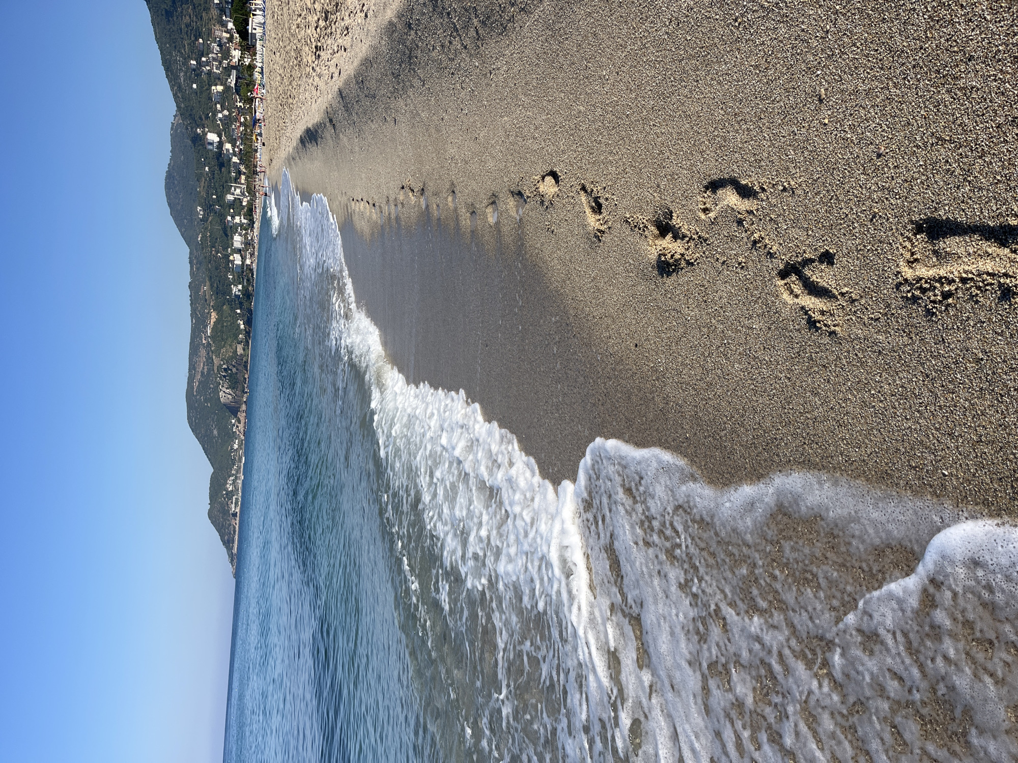 This image captures a picturesque coastal scene in Alanya, Turkey, on a bright, sunny day. The perspective is low, looking along the shoreline where the clear blue sea meets a sandy beach.
In the foreground, a textured sandy beach extends diagonally across the frame. Numerous distinct footprints are impressed into the damp sand near the water's edge, suggesting a recent stroll. Gentle waves with white, foamy crests are breaking and receding onto the shore, creating a dynamic edge between the land and the sea. The water is a serene blue, transitioning from the frothy white closer to the beach to a smooth surface further out.
The midground features the expansive, calm sea, stretching towards a distant horizon. Along the far coastline, a range of large, green, tree-covered mountains rises steeply. Numerous white and light-colored buildings are visible nestled along the mountain slopes and at their base, indicating a developed coastal town or resort area. A few very small, indistinct figures can be observed on the distant beach, adding a subtle human presence.
The sky above is a brilliant, cloudless blue, filling the upper portion of the scene. The overall impression is one of a peaceful and inviting seaside landscape under clear, warm weather. No visible text is present in the image.