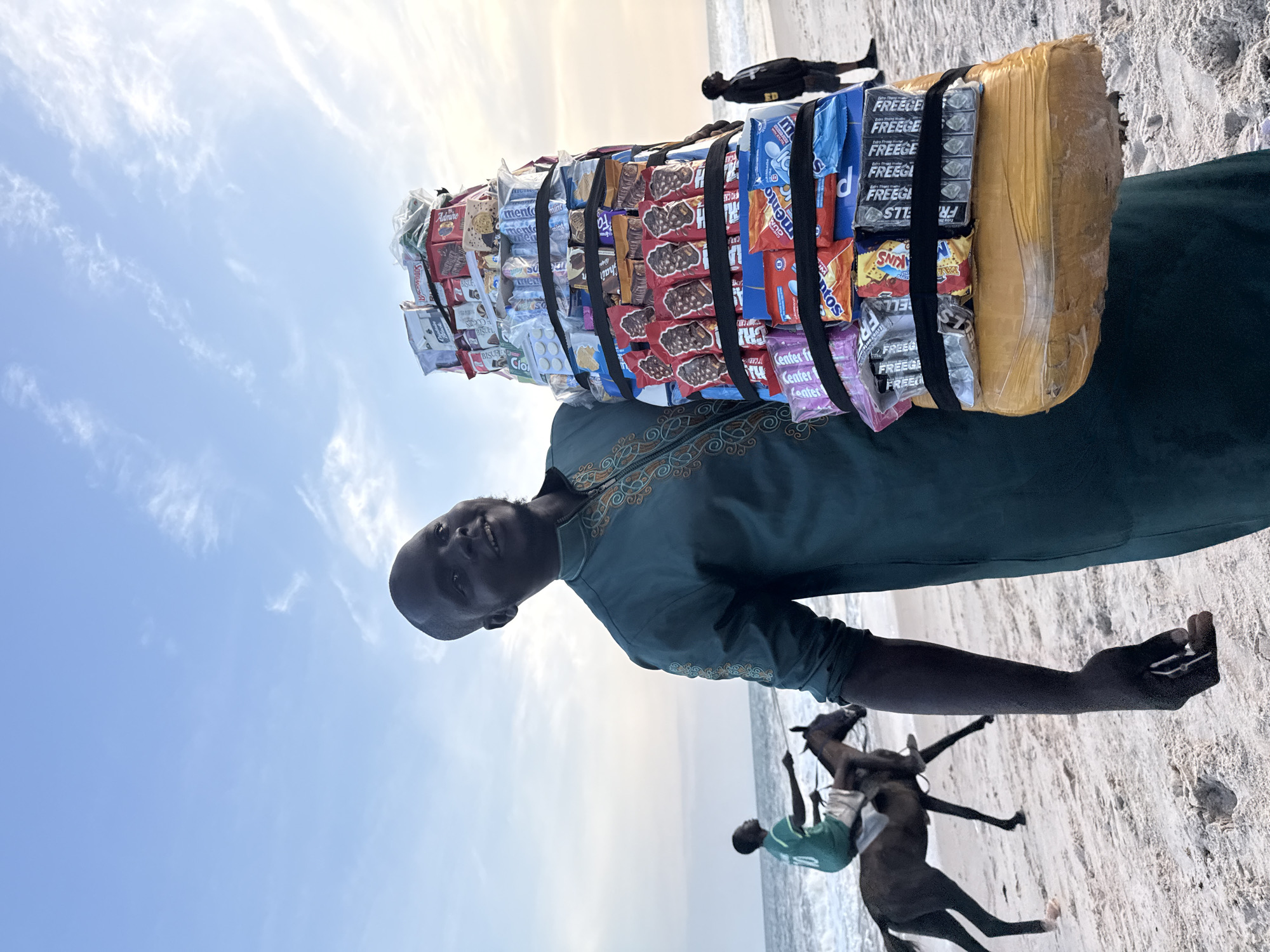 A man stands on a sandy beach, carrying a large stack of candies and gum on his head. He is facing towards the viewer with a smile. He wears a green, patterned shirt. Behind him, near the water's edge, are two people on horseback. The sky above is light blue with wispy clouds. The image is possibly taken in Mopo Onijebu, Nigeria. The candies appear to be secured with black straps and wrapped in yellow packaging. There are brands such as "Mentos," "Center Fresh," and "Freege" visible.