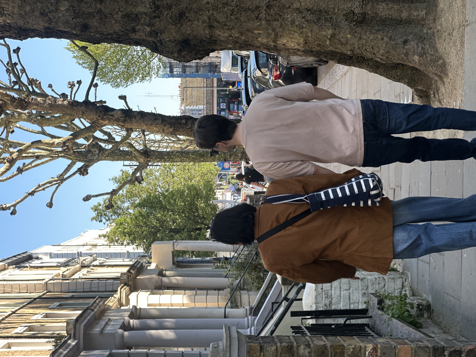 Two people walk along a residential street in London, United Kingdom. The buildings are traditional row houses with light-colored facades and classical architectural details. A large tree canopy extends over the street, partially obscuring the sky. Traffic and shops are visible in the distance. The person in the foreground wears a brown jacket and blue jeans, carrying a black and white striped bag over their shoulder. The person behind them wears a pale pink shirt and blue jeans. The perspective is from street level. The image appears to be rotated 90 degrees counter-clockwise.