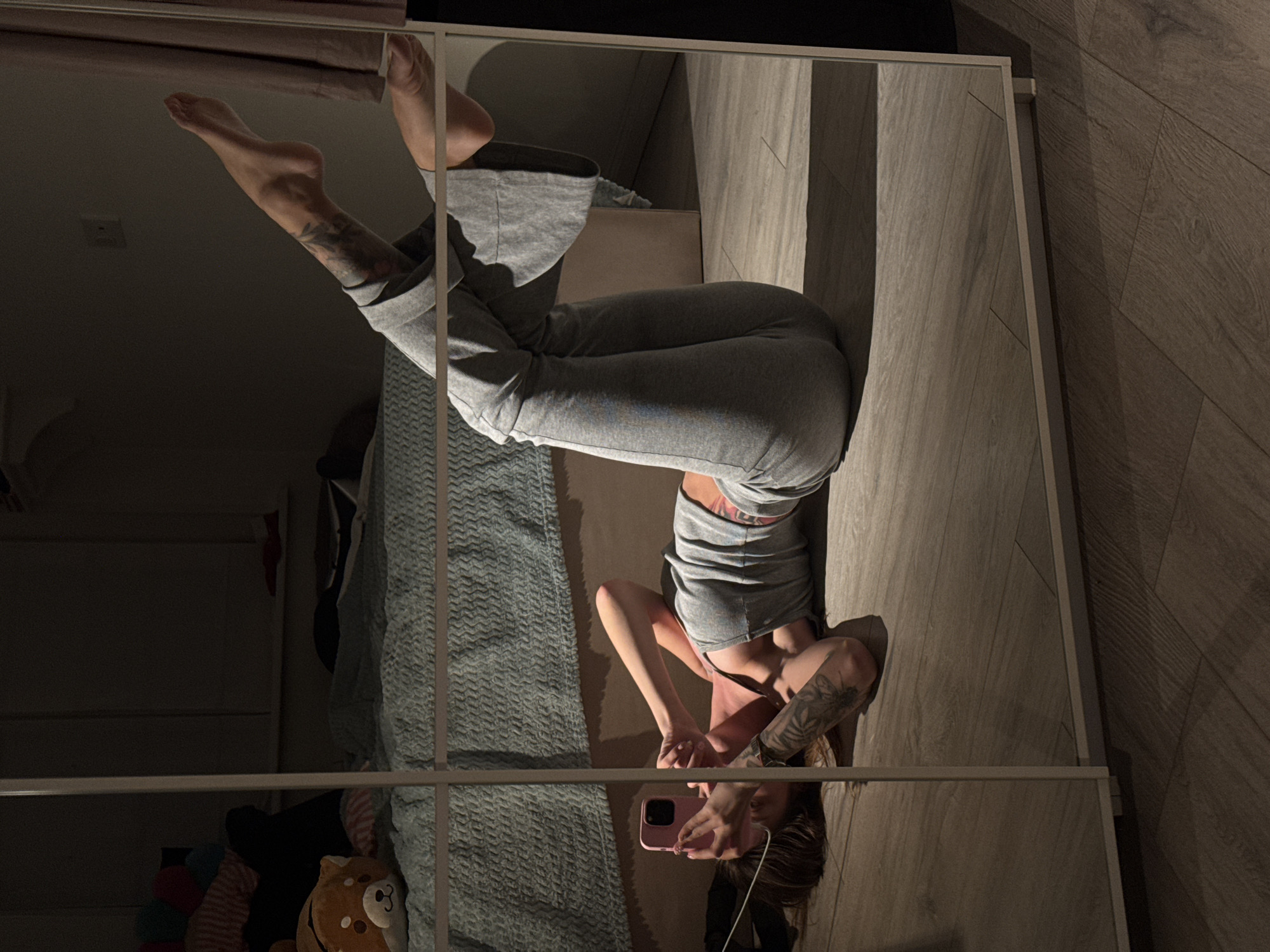 The image depicts a young woman lying on her back on a wooden floor, posing for a selfie in a full-length mirror. She is wearing a grey tank top and matching grey flared pants. Her legs are bent at the knees, and her feet are raised. She is holding a pink smartphone in her left hand, with her right arm resting above her head. Her hair is dark and falls over her shoulders. The background, reflected in the mirror, shows a bedroom with a bed covered in a textured blue blanket and several stuffed animals on it. The lighting in the room is dim, with a strong light source casting shadows on the floor and walls. There are no visible indications of the city of Moscow, Russia.
