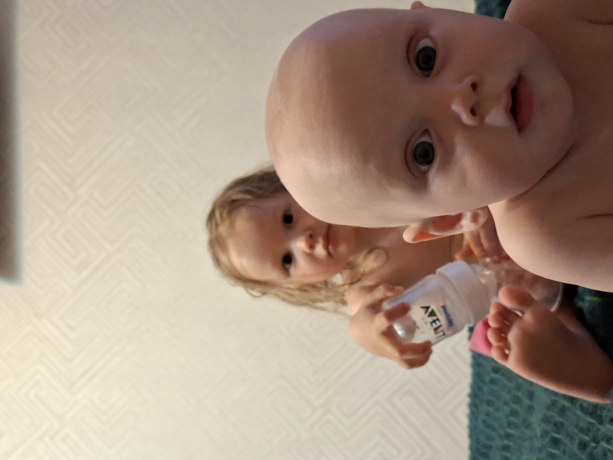 The image shows two children, one infant and one slightly older child, in an indoor setting. The infant, with a round head and a surprised expression, is in the foreground. The older child is behind, holding an Avent baby bottle. The background features a wall with a geometric pattern and a blue textured surface.