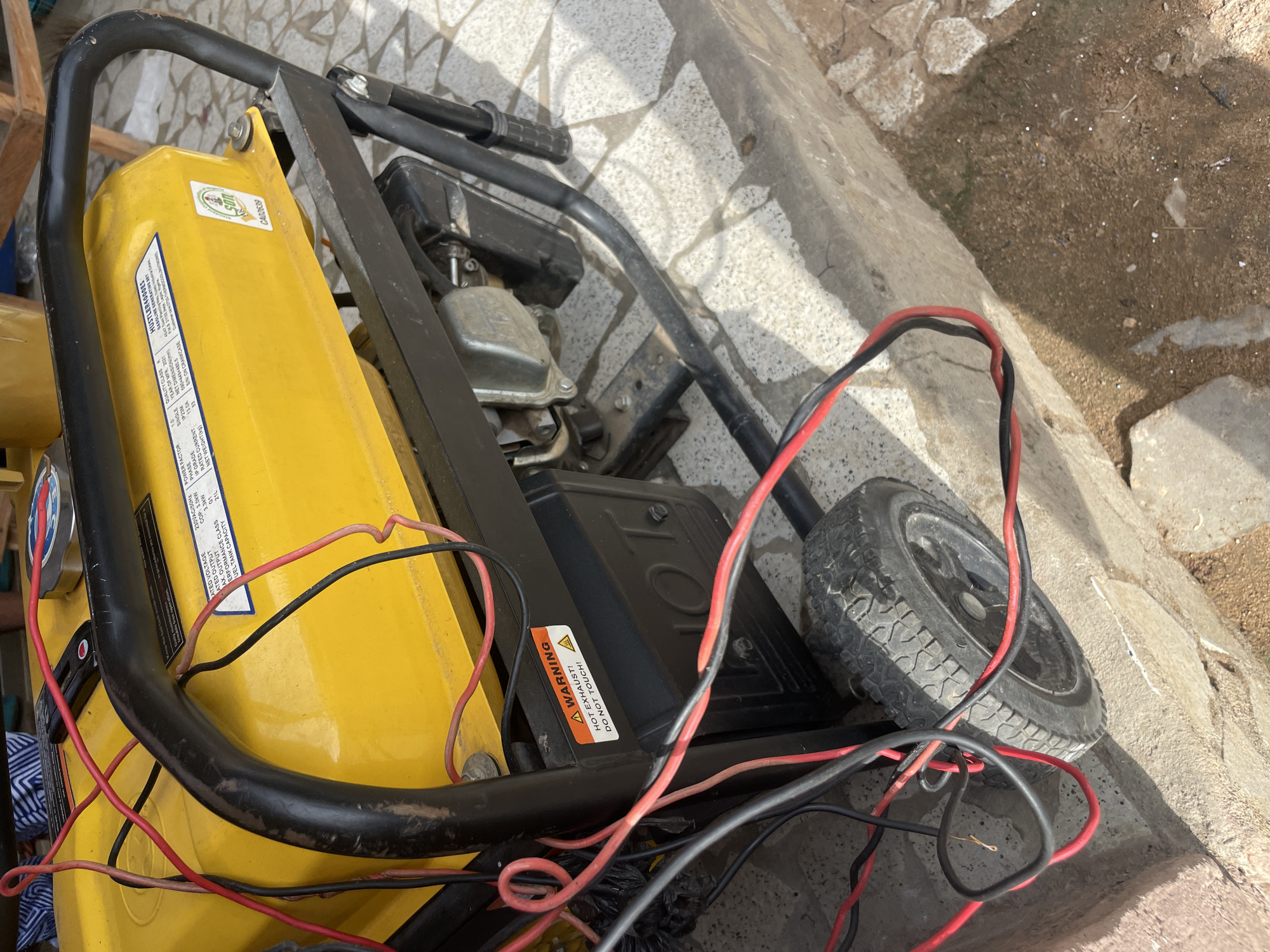 The image shows a yellow portable generator with black metal framing and a black handle bar. Red and black electrical wires are connected to the generator and are draped across its surface and the surrounding environment. The generator is positioned on a concrete surface, possibly a sidewalk or step, with rough, sandy ground visible in the lower right portion of the frame. A black tire is attached to the left side of the generator. There is a warning sticker on the generator that reads "A WARNING HOT EXHAUST! DO NOT TOUCH!". The scene appears to be outdoors.