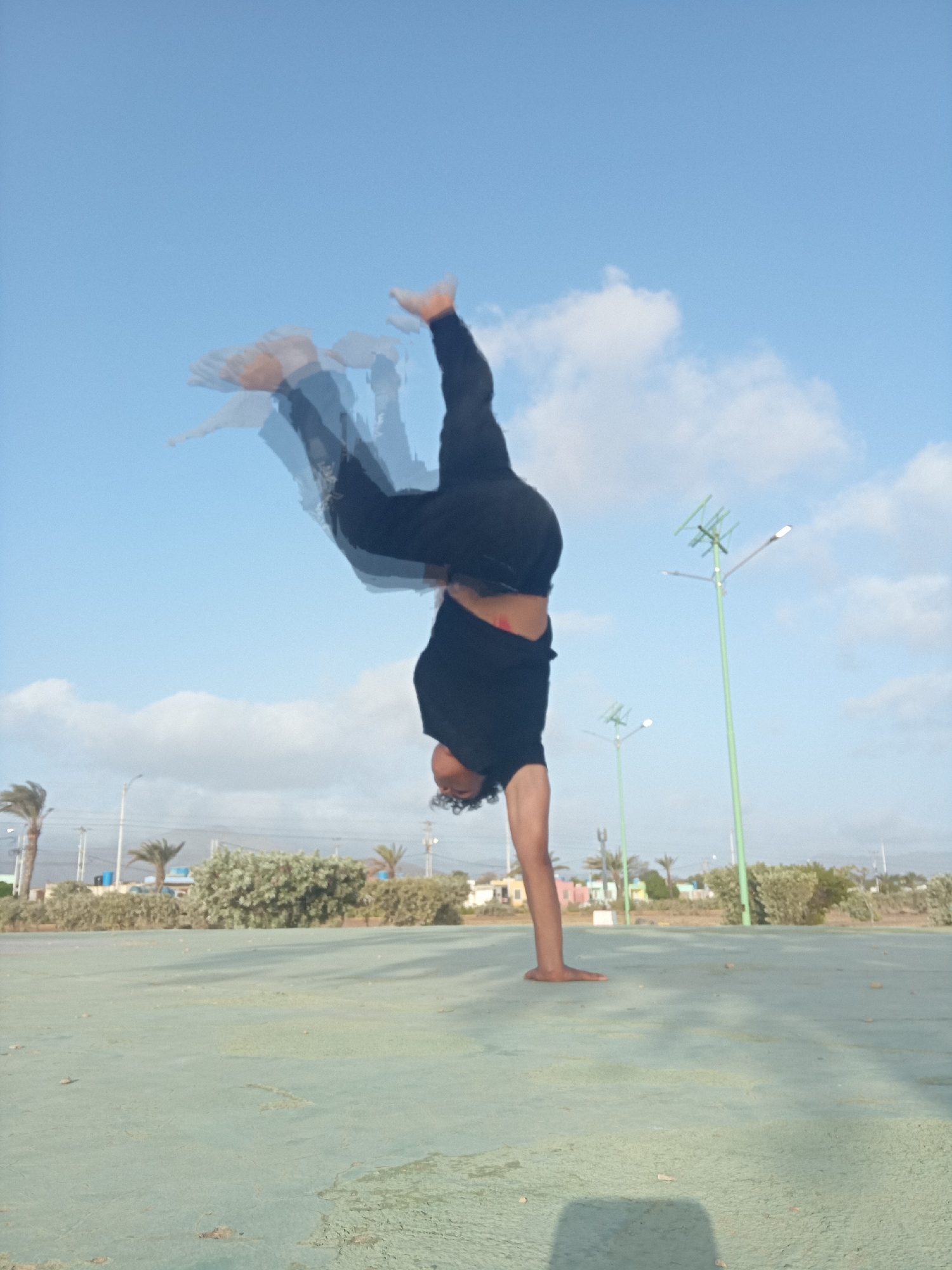An outdoor, eye-level shot captures a person performing an acrobatic maneuver, likely a handstand or a cartwheel, in an open area during daytime. The individual, dressed in a black t-shirt and black pants, is depicted upside down, balancing on one hand. A prominent motion blur effect is visible across their body, especially in the legs, conveying the dynamic nature of the movement. Their head, with curly dark hair, is visible near the ground.
The setting features a light green, possibly paved or concrete, ground surface that stretches into the foreground. The background is dominated by a clear blue sky with scattered white clouds. Several green streetlights or lampposts with modern fixtures stand tall against the sky, with one prominent on the right. A line of green bushes and trees, including several palm trees, defines the horizon. Some palm trees appear wind-blown, suggesting breezy conditions. Faint structures or buildings are discernible in the far background. The bright natural lighting indicates it is daytime, possibly late morning or afternoon, consistent with the sunny environment of Juan Griego, Venezuela. No text is visible in the image.