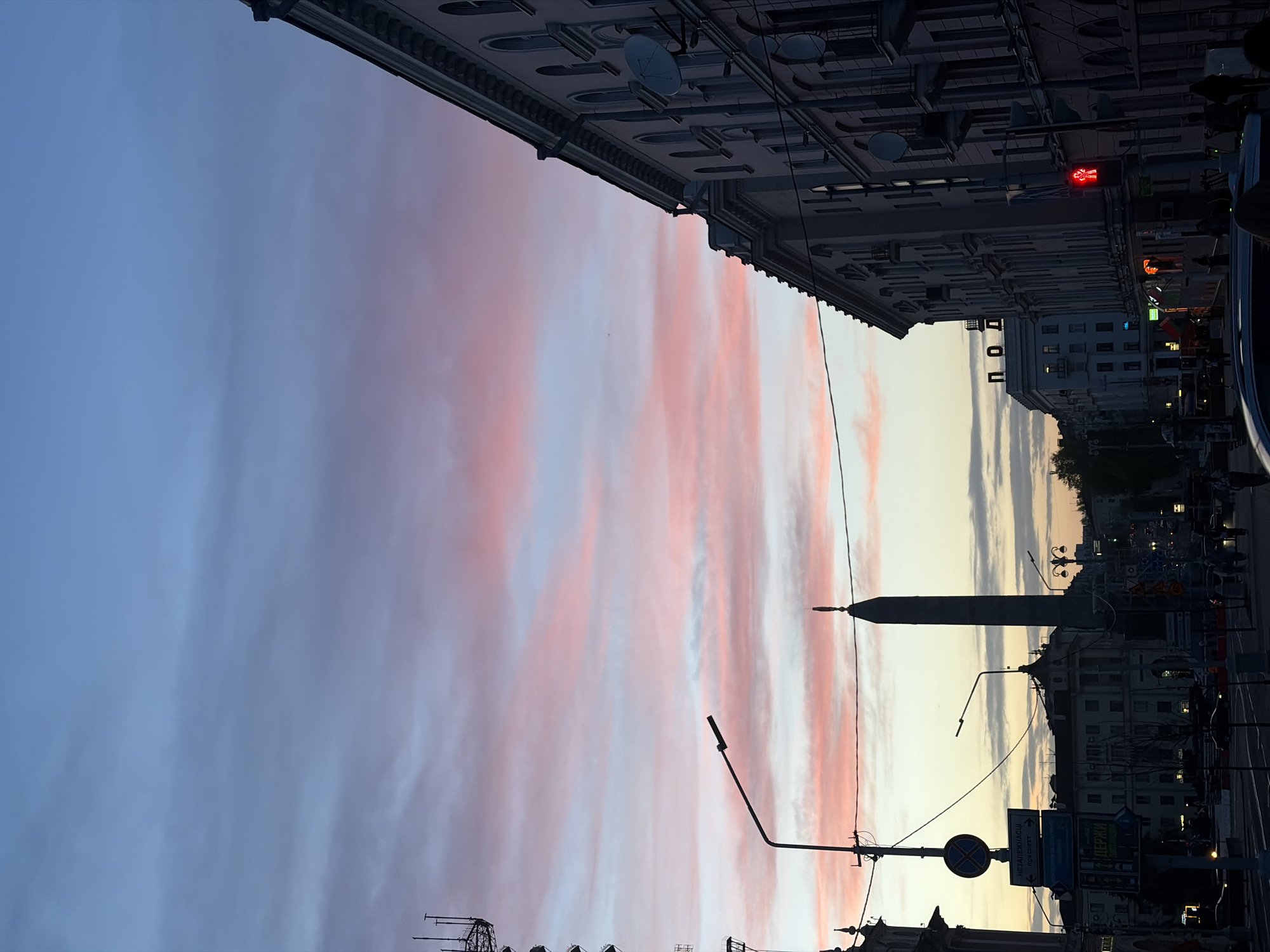 The image shows a street scene in Minsk, Belarus, during twilight. The sky is filled with clouds tinted pink and purple from the setting sun. On either side of the sky, there are buildings. The building on the upper left is taller with detailed architectural features and satellite dishes. The building on the right is illuminated with lit windows and street lights, including a red traffic light. In the center of the image is Victory Square with the Victory Monument. There are lampposts and overhead wires that stretch across the view. In the foreground, there are street signs and silhouettes of other buildings and structures.