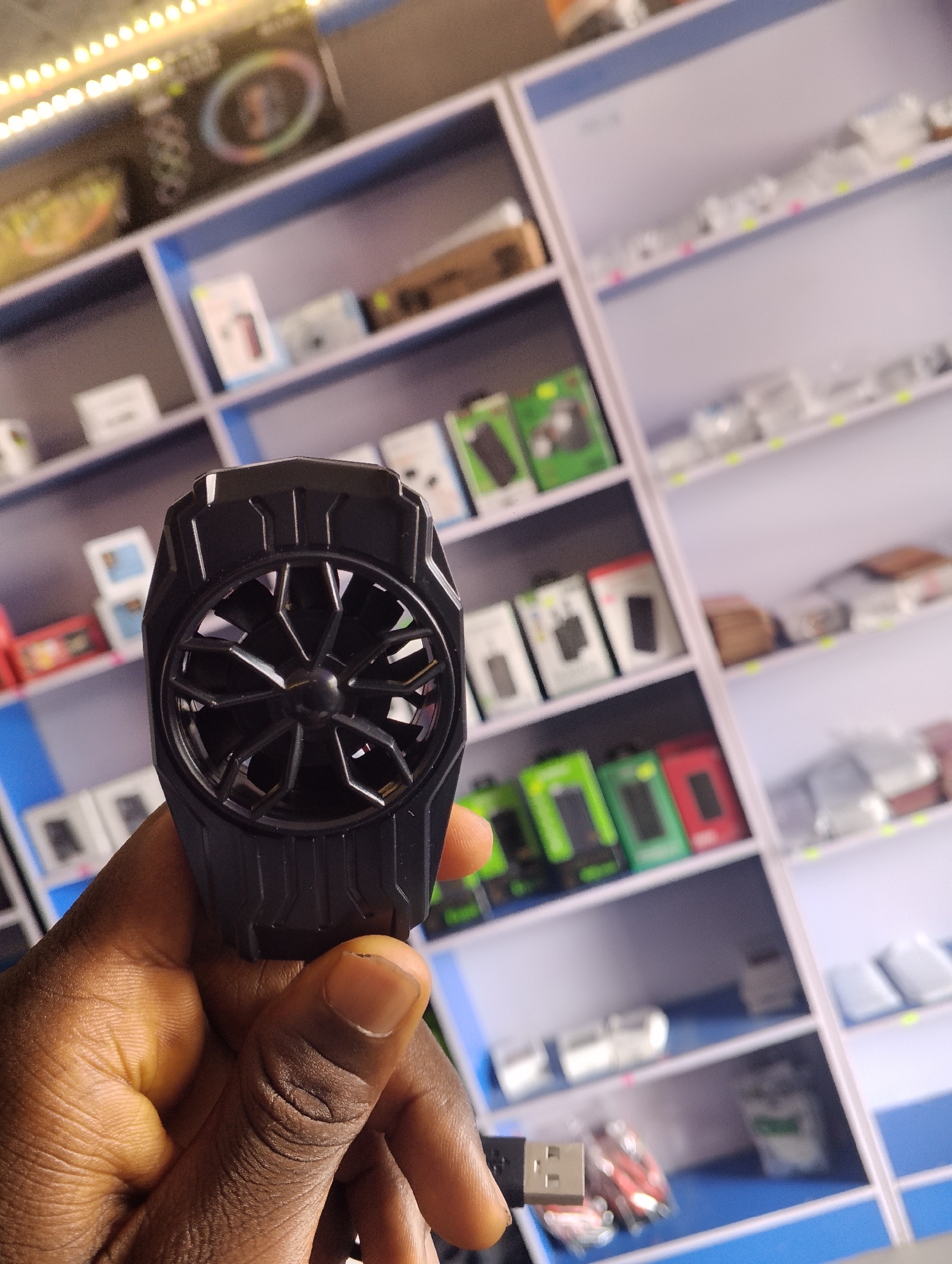 A close-up shot shows a person's hand holding a black, fan-like device, possibly a phone cooler, in front of shelves stocked with electronics. The device has a circular grill with fan blades visible inside. A USB cable extends from the bottom of the device, with its connector visible. The background consists of white shelves with blue trim, filled with various packaged electronic items, some in green, white, and red boxes. The overall lighting suggests an indoor retail environment, consistent with a shop in Jalingo, Nigeria. The focus is on the device and the hand, with the background being out of focus.
