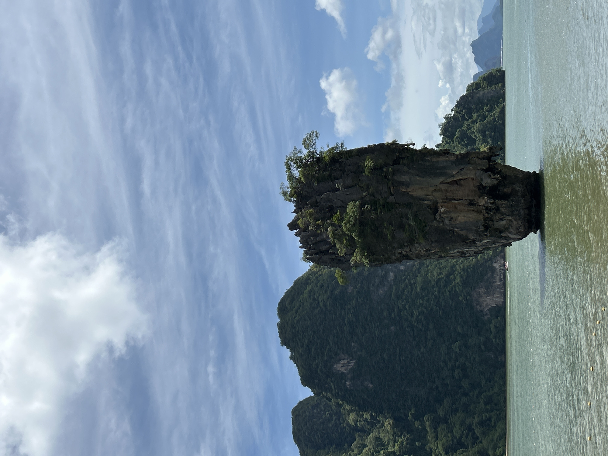 The image depicts a tall, slender limestone karst in the middle of a body of water, likely the Andaman Sea. The karst is covered in lush green vegetation, with trees growing from its crevices. In the background, several larger, jungle-covered islands rise from the sea, their peaks shrouded in mist. The sky is a bright blue with scattered white clouds. The water is a calm, greenish hue, reflecting the surrounding landscape. A small boat is visible in the distance, moving across the water. The scene is serene and picturesque, characteristic of the scenery found in Phang Nga Bay, near the city of Krasom, Thailand.
