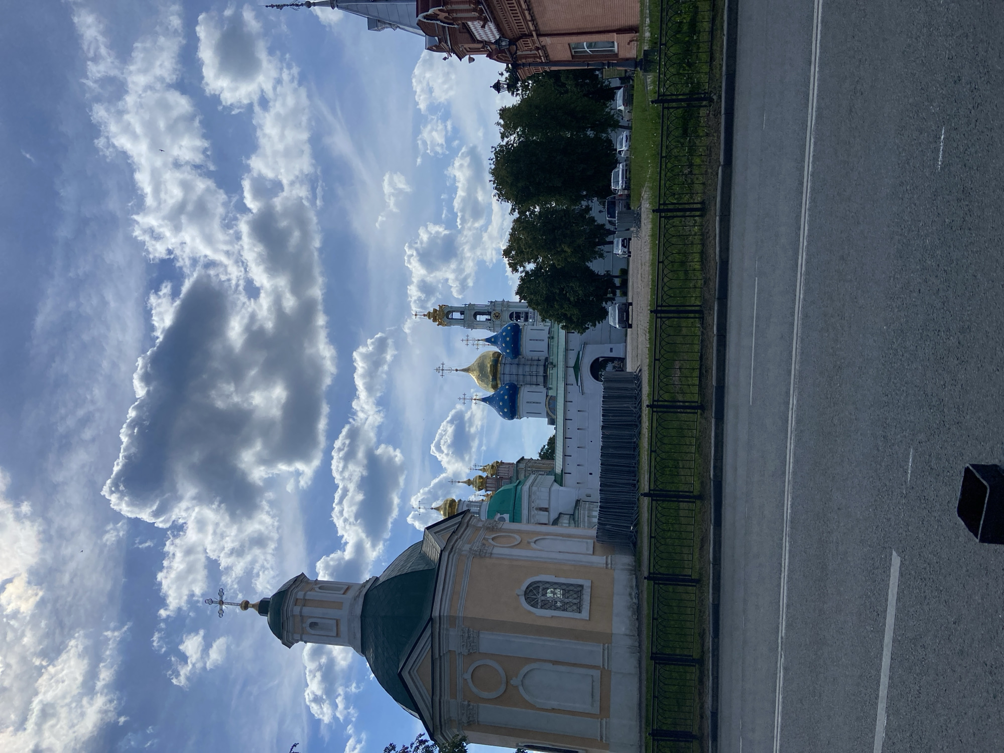 This outdoor image captures a picturesque architectural landscape in Sergiyev Posad, Russia, showcasing elements of the Trinity Lavra of St. Sergius.
In the foreground, an empty asphalt road with white lane markings stretches horizontally, separated from the main complex by a black wrought-iron fence and a strip of green grass. Beyond the fence, a line of lush green trees partially obscures the view of parked cars and other buildings, including one with red brickwork.
The central subjects are several magnificent Russian Orthodox churches and cathedrals. On the left, a light yellow building with a dark green dome topped by a golden cross stands prominently. Further into the complex, a tall bell tower with white walls and a golden-domed top is visible, flanked by other religious structures. These buildings feature distinctive onion domes in various colors, including bright gold, deep blue, and green, each adorned with Orthodox crosses. The intricate details of the architecture, such as arched windows and decorative elements, are visible.
The scene unfolds under a bright blue sky, which is filled with an array of dramatic white and grey cumulus clouds, indicating a clear, sunny day. A small bird is visible soaring high in the upper left portion of the sky.
The overall atmosphere conveys a sense of historical grandeur and serenity. No people or discernible text are visible in the image. The time of day is clearly daylight, likely midday or early afternoon, given the bright illumination.
