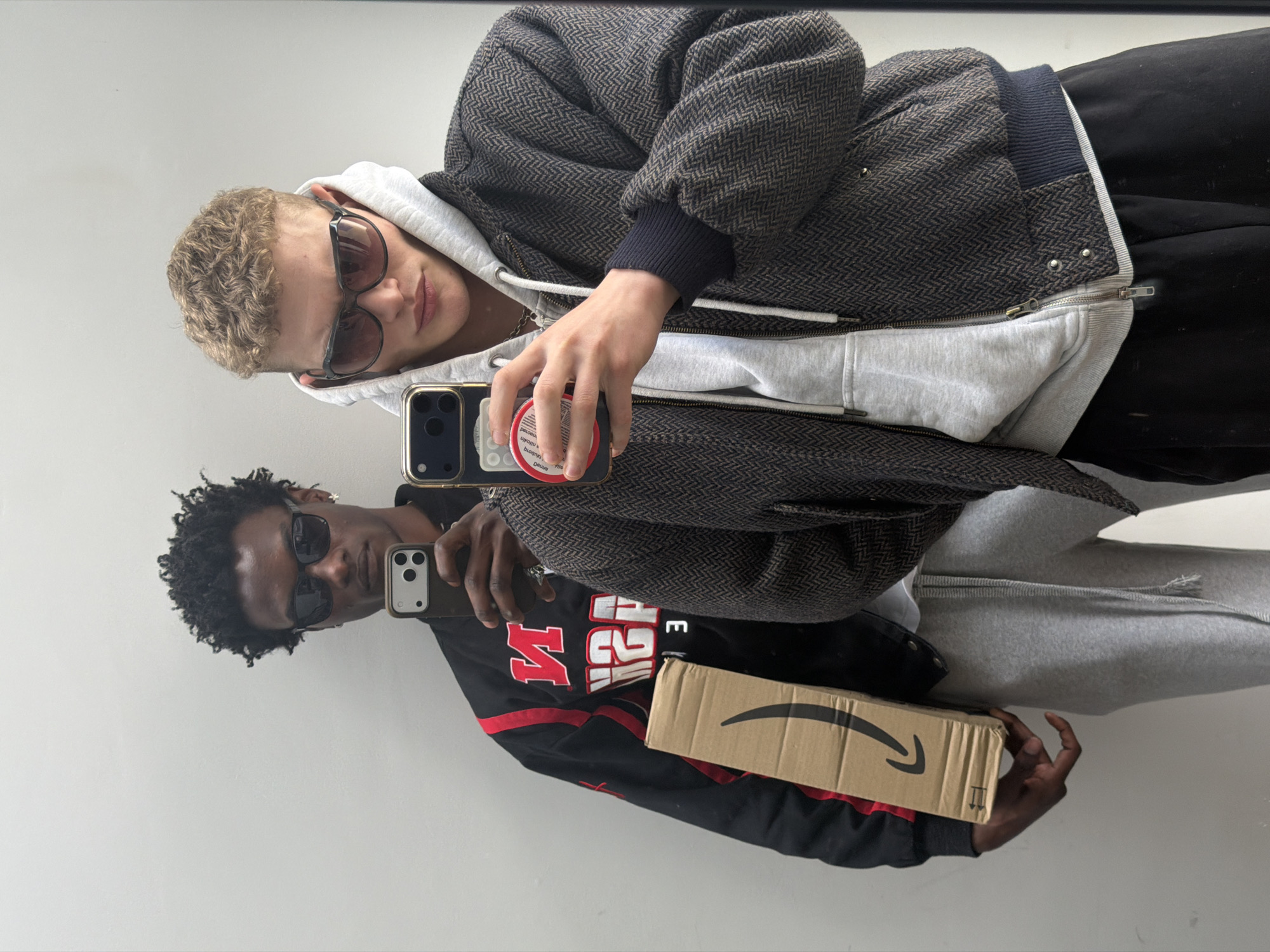 Two individuals are visible in a mirror selfie. One person, presumably of Caucasian descent, is wearing sunglasses, a white hoodie, and a patterned jacket. He holds a smartphone in his right hand. The second person, with dark skin, is wearing sunglasses and a black and red jersey. He holds a smartphone and an Amazon box. The background is a plain white wall. The location cannot be determined from this image.
