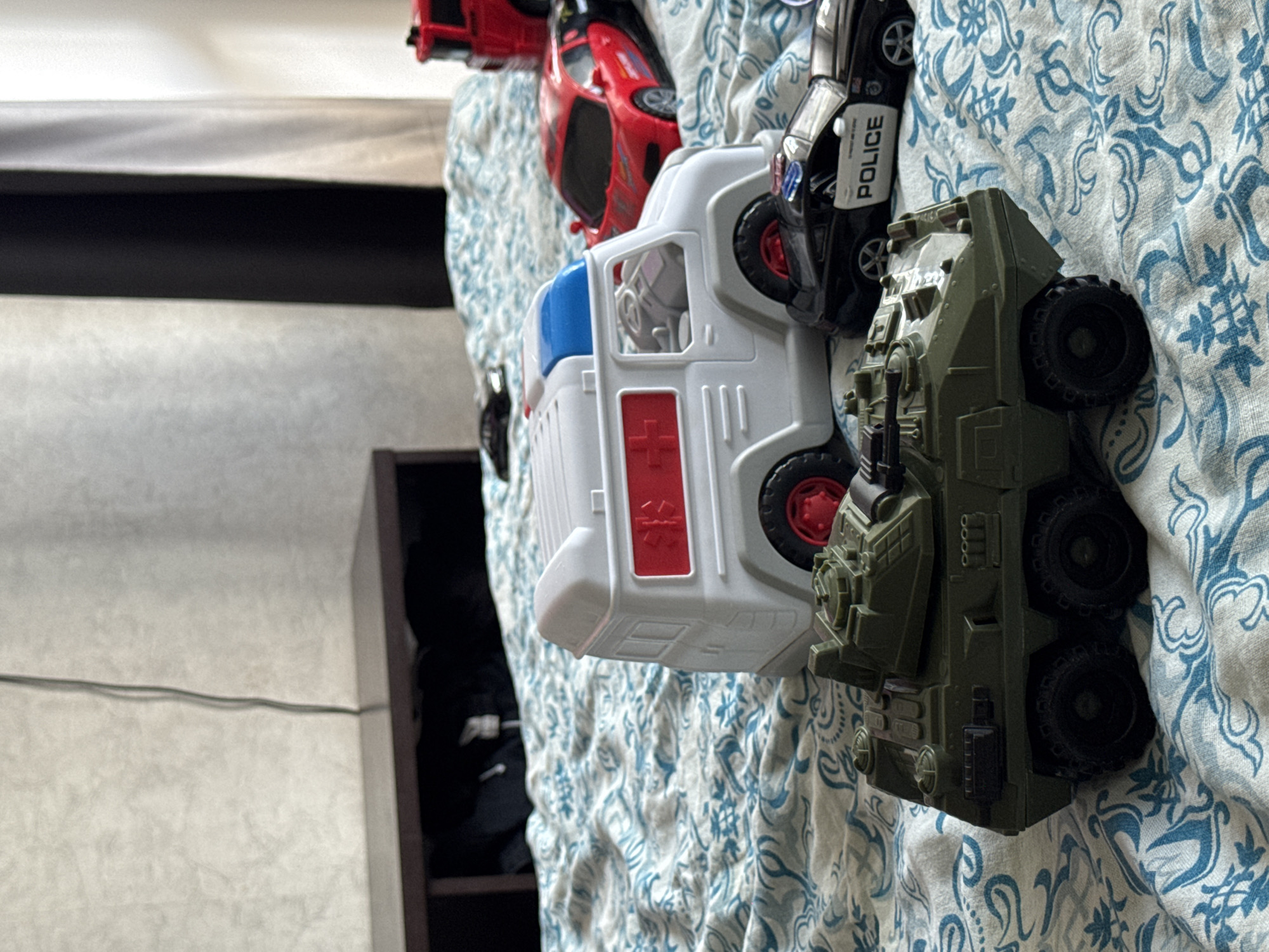 The image displays a collection of toy vehicles arranged on a patterned fabric, likely a bedspread or blanket, with a light blue and white floral design. The toys are lined up horizontally across the frame.
The subjects are toy vehicles:
- A red toy sports car is partially visible on the left.
- A white toy ambulance with red accents, including a cross symbol and blue lights on top, is prominent in the center.
- A black toy police car with "POLICE" written in white on its side is next to the ambulance.
- A green toy military vehicle with a rugged design and large wheels is on the right.
The scene is static, with the toys simply placed on the fabric. There is no discernible activity or event taking place.
Notable details include the textured pattern of the fabric and the realistic details of the toy vehicles, such as wheels, windows, and markings. The lighting appears to be natural, suggesting daytime. The setting is a domestic indoor space, as indicated by the fabric and the edge of a shelf or furniture visible on the left.
The visible text on the toys includes "POLICE" on the black car. There is no text related to the location context provided.