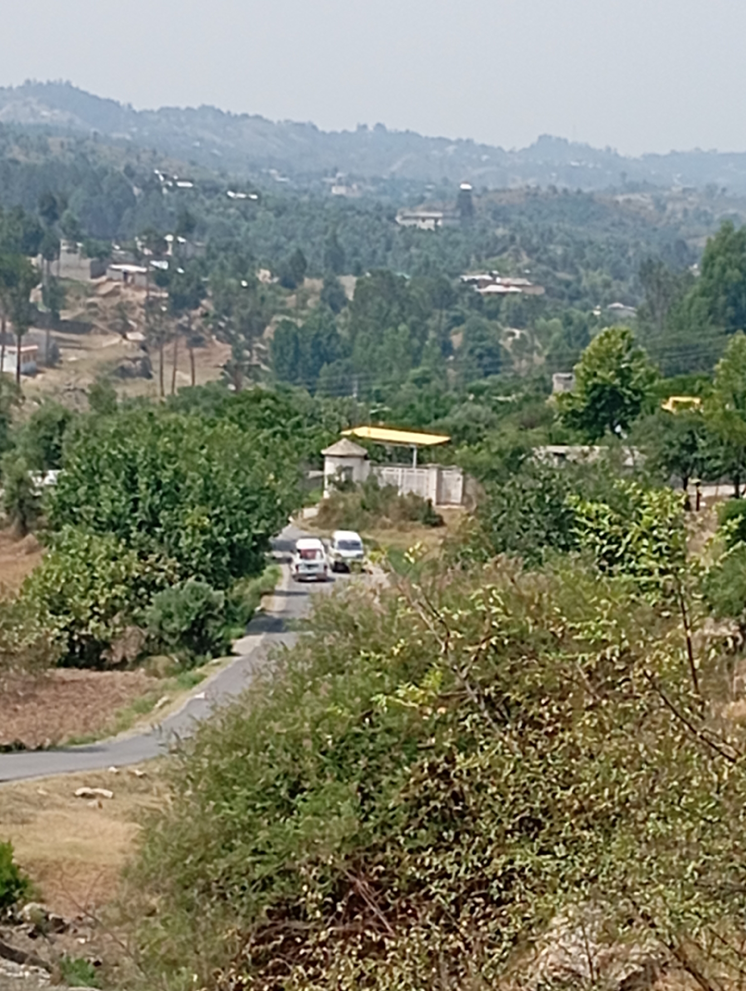 The image shows a rural landscape in the Hakale region of Pakistan, characterized by rolling hills covered in dense green foliage. A paved road winds through the scene, with two white vans driving on it. One van is closer to the foreground, while the other is further down the road. On the right side of the road, there is a building with a yellow roof and white walls, appearing to be a bus stop or a small shelter. In the background, more hills and scattered houses are visible, indicative of a village or small town nestled within the natural landscape. The sky is overcast, suggesting a hazy or cloudy day. The foreground is dominated by lush green bushes and trees, creating a natural framing for the road and vehicles. The overall impression is one of a tranquil, remote environment typical of the Pakistani countryside.