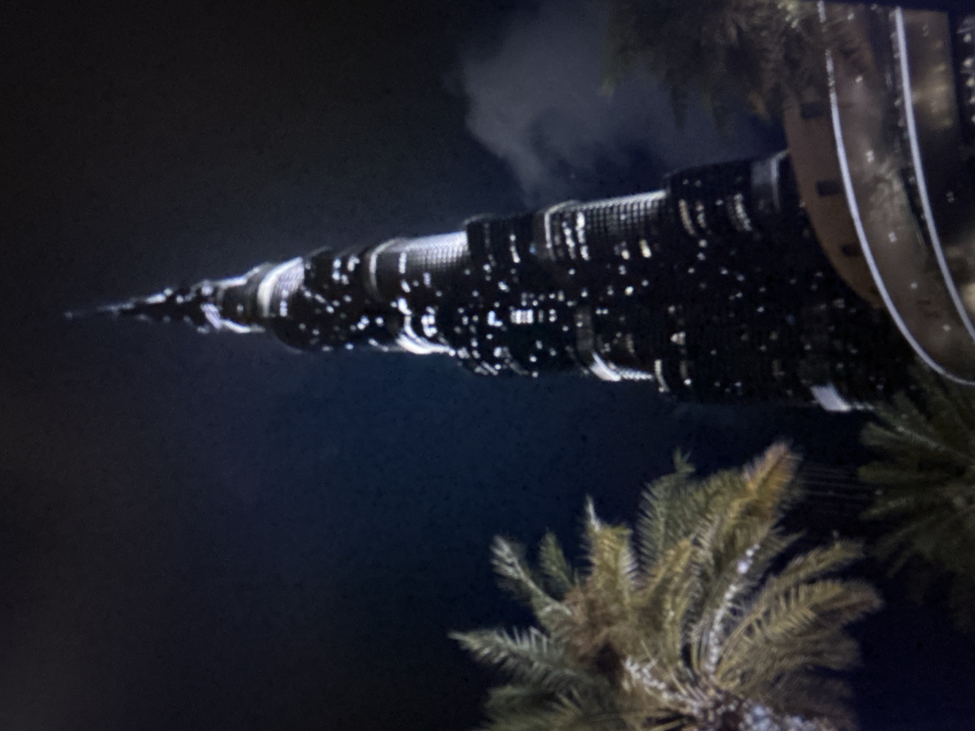 The image depicts the Burj Khalifa in Dubai, United Arab Emirates, photographed at night. This scene is not located in Burgas, Bulgaria. The central subject is the exceptionally tall, illuminated skyscraper, which tapers distinctly towards a spire at its apex. Numerous lights are visible across the dark facade of the building, creating a speckled pattern. In the left foreground, several palm trees with discernible fronds are visible, silhouetted against the dark night sky. The sky itself is a deep blue-black, with subtle cloud formations present in the upper right quadrant. A curved, multi-tiered architectural element with embedded lights is partially visible at the base of the skyscraper. No individuals are present in the frame.
