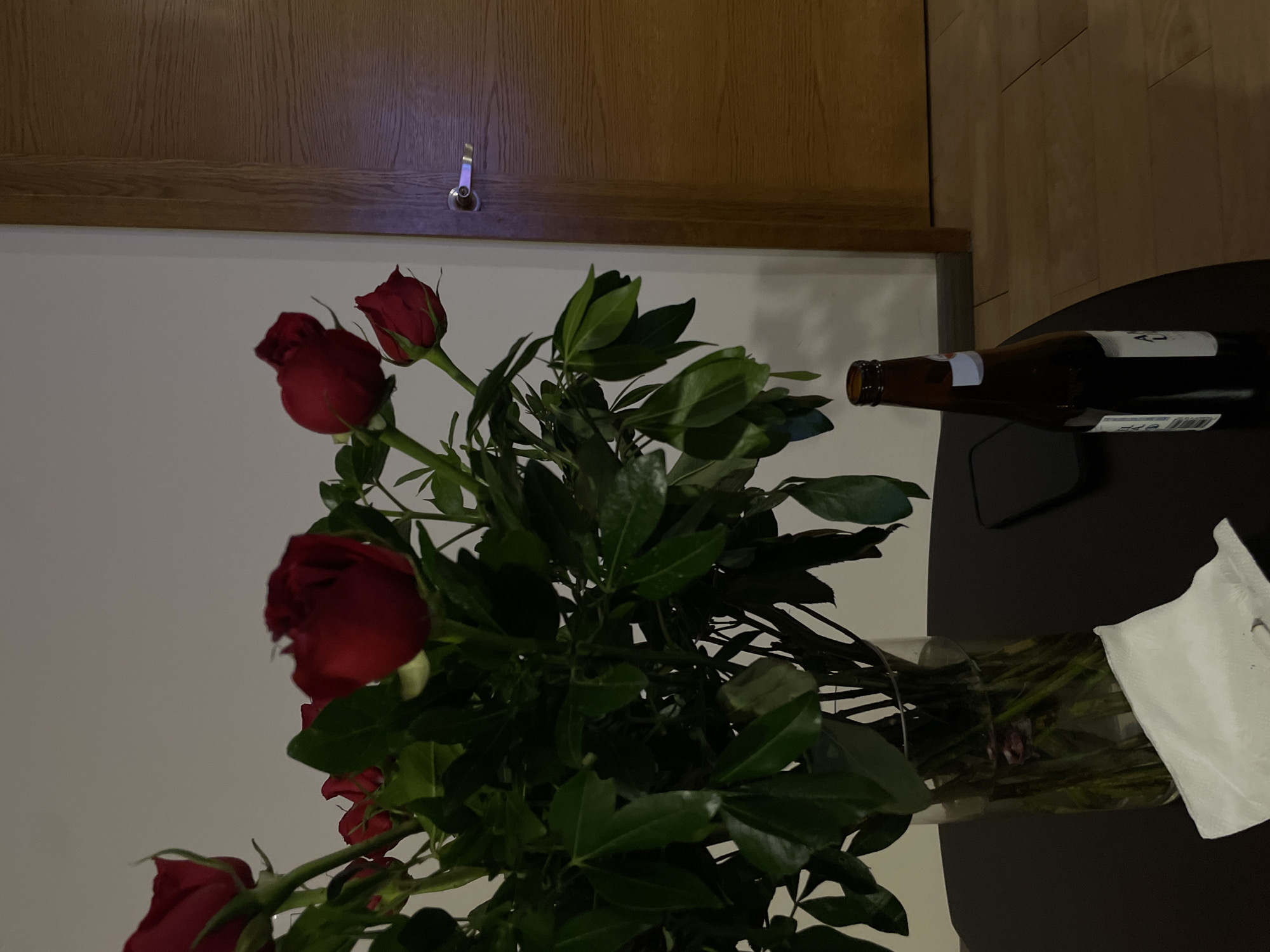 The image shows a vase of red roses on a dark surface, likely a table. A beer bottle and a white napkin are also present on the table. The background includes a white wall and a wooden cabinet. The scene appears to be indoors, possibly in a private residence. There is no information in the picture to indicate this is in Puebla, Mexico.