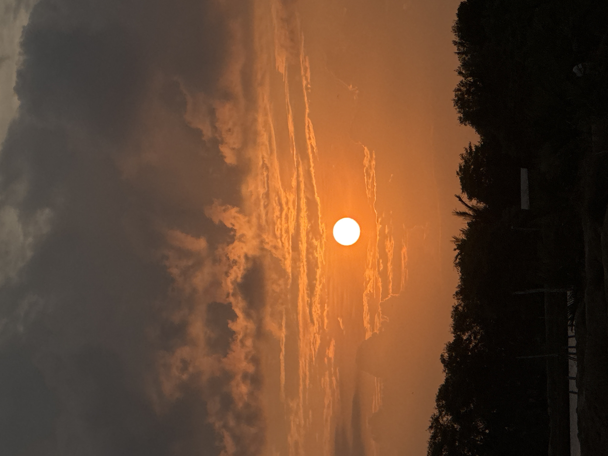 The image depicts a sunset with a prominent, bright sun partially obscured by clouds. The sky is dominated by shades of orange and dark gray, with illuminated clouds forming distinct patterns. In the foreground, a dark silhouette of trees spans the lower portion of the image, with some indistinct structures and possibly faint figures visible at the very bottom. The scene appears to be set in an outdoor environment, likely rural, given the dense tree line. The specific location in Mesjid Ulim Baroh, Indonesia, suggests a tropical or sub-tropical setting, consistent with the observed flora. The lighting indicates it is late evening.