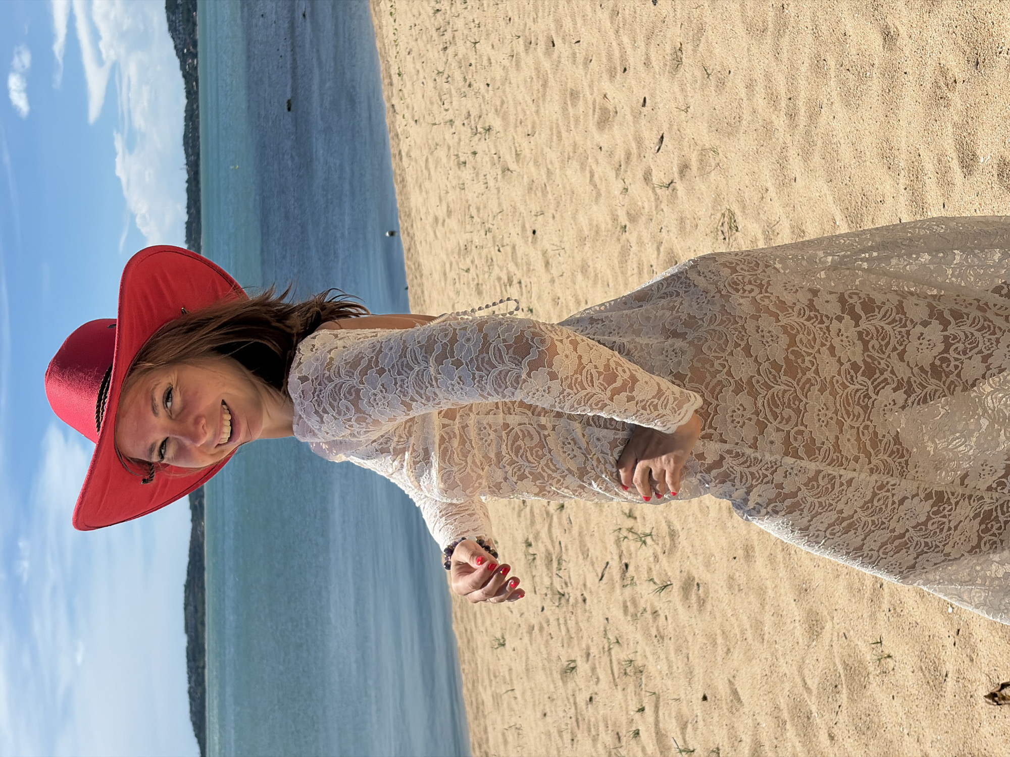 A woman wearing a white lace dress and a red hat stands on a sandy beach in Baan Bo Phut, Thailand. The ocean is visible in the background, with a hilly coastline in the distance under a blue sky with scattered clouds. The woman is smiling and looking over her shoulder at the camera, with her right hand on her hip and her left hand down by her side, wearing a bracelet. The sand is light brown and has sparse patches of dry grass.