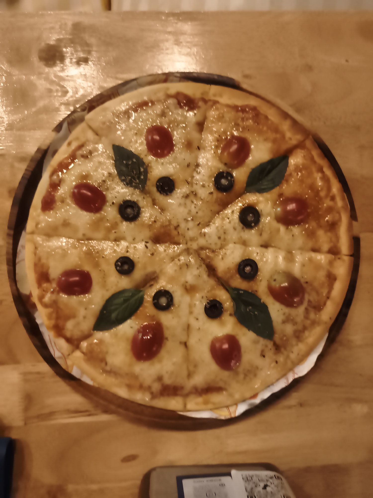 A pizza is presented on a wooden table. The pizza is round, sliced into eight pieces, and topped with melted cheese, cherry tomatoes, black olives, and basil leaves. The scene appears to be indoors, likely a restaurant, given the wooden table and the pizza's presentation. The lighting suggests it could be evening or an indoor setting with artificial lights. There are no people visible in the image, and no specific weather conditions can be inferred. The location context provided is "บ้านบางรักษ์, Thailand," which might indicate the general area where the photo was taken. There is some visible text on a card or device in the lower part of the image, but it is not clear enough to be read.