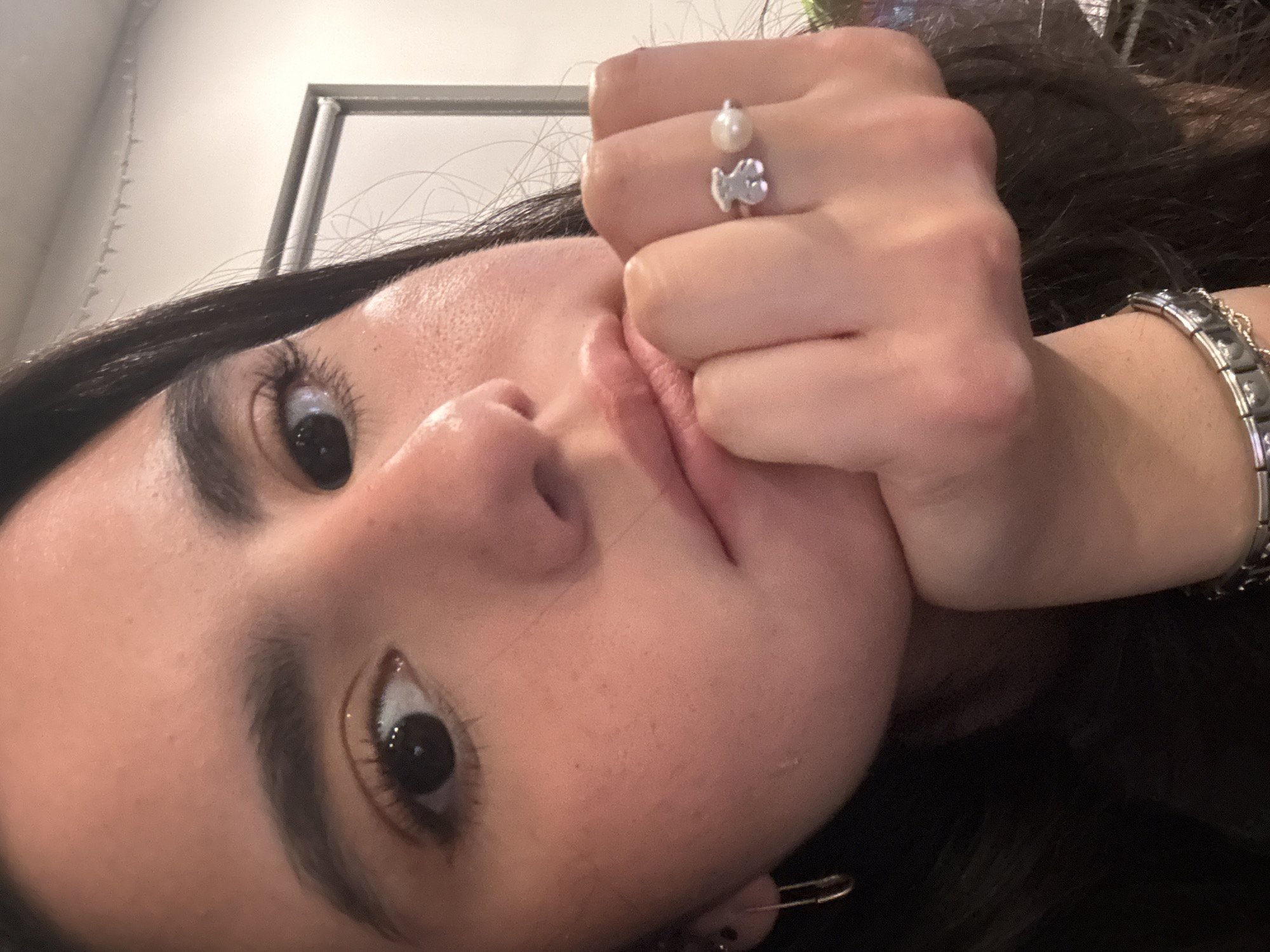 A close-up, slightly rotated, indoor photograph features a young woman with dark hair and brown eyes. Her face is the primary subject, with her left hand clenched around her face near her mouth. The fingers of her hand display a ring with a pearl and a bear-shaped charm. On her right wrist, a silver-toned bracelet is visible. A gold safety pin is seen near her ear. The background includes a white wall and a framed object out of focus in the upper portion of the frame. Without additional context or metadata in the image, I cannot determine whether this photograph was taken in the city of Austin, United States of America.