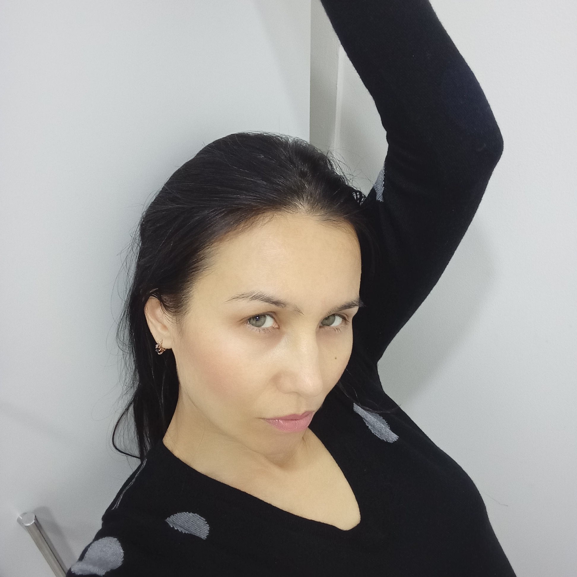 A medium close-up portrait shows a woman indoors, possibly in a residential setting. She has dark hair pulled back, fair skin, and light-colored eyes. She wears a black long-sleeved shirt with gray polka dot-like patterns. Her right arm is raised and slightly bent at the elbow, leaning against a light gray or white wall. The background is a plain, undecorated wall. A metallic, cylindrical object is faintly visible on the lower left side of the frame. The location is not identifiable as Izhevsk, Russia, or any other specific location from the visual information alone.