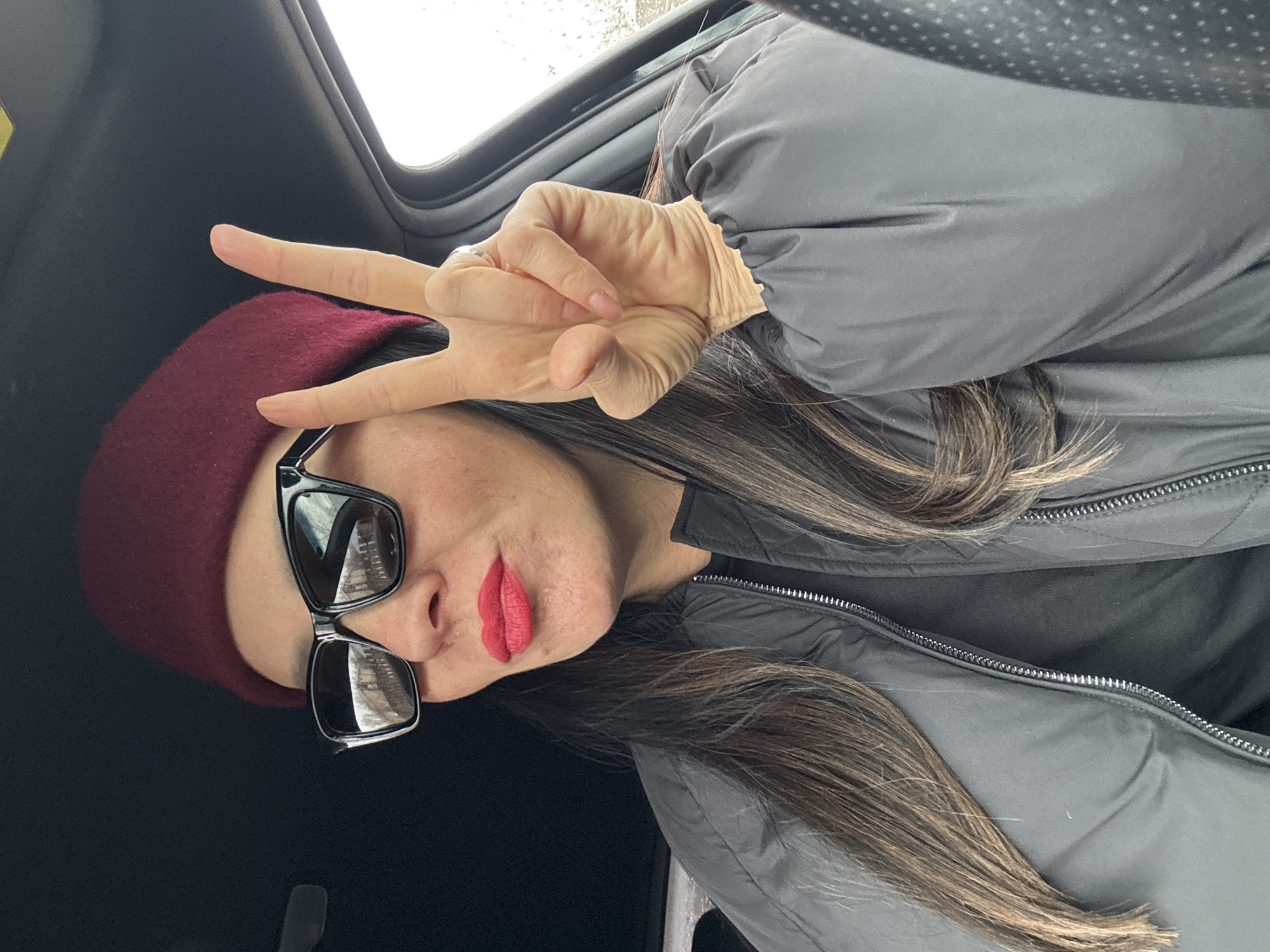 A woman is pictured indoors, likely within a vehicle due to the sunroof visible in the upper portion of the frame. The woman is wearing a maroon knit cap, black sunglasses, and a gray quilted jacket. She has long, dark hair with highlights, and she is wearing red lipstick. Her right hand is raised, making a peace sign. The location cannot be determined to be within the city of Chaikovsky, Russia based solely on the visual information provided.