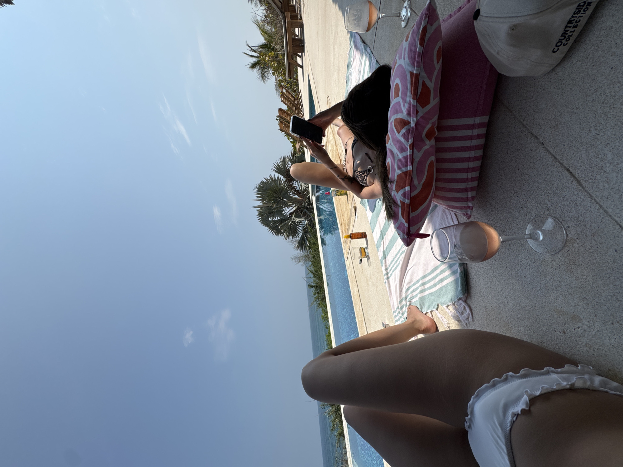 A poolside scene is captured, with two individuals in different states of activity. One person, positioned against a pink pillow, lies on a striped towel and is engrossed in their phone, their swimsuit visible. Nearby are two wine glasses. Another individual, wearing a white bikini, is situated closer to the viewer and appears to be sunbathing. The pool itself is visible, flanked by lush greenery, possibly palm trees, hinting at a tropical location. A portion of a building structure is seen in the background. The location is Unknown, Mexico.