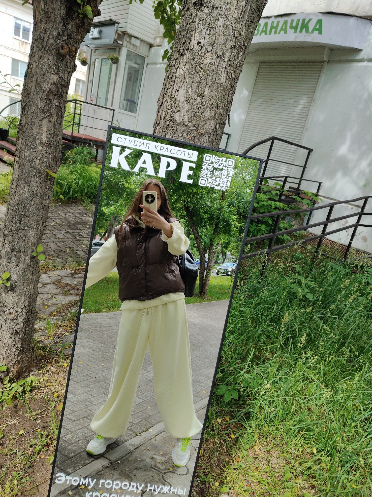 An outdoor, eye-level shot captures a woman taking a mirror selfie on a paved path. The woman, reflected in a large vertical mirror, wears a brown puffer vest over a light yellow long-sleeved top and matching wide-leg pants, paired with white sneakers accented with neon green. She holds a light-colored smartphone, obscuring most of her face, to capture her reflection. A black backpack is visible on her left shoulder.
The mirror itself is bordered in black and prominently displays white text at the top: "СТУДИЯ КРАСОТЫ" (Beauty Studio) and "КАРЕ" (KARE), flanked by two QR codes. At the bottom, partially cut off, are the words "ЭТОМУ ГОРОДУ НУЖНЫ" (This city needs).
The scene is bathed in bright daylight. The mirror is positioned between two trees with rough bark. On the left, behind the mirror, is a light-colored building with windows and an entrance staircase. On the right, another white building features a closed metal shutter and a green sign with the word "ЗАНАЧКА" (ZANACHKA). Lush green grass and foliage are visible in the foreground and background, indicating an urban or suburban setting.