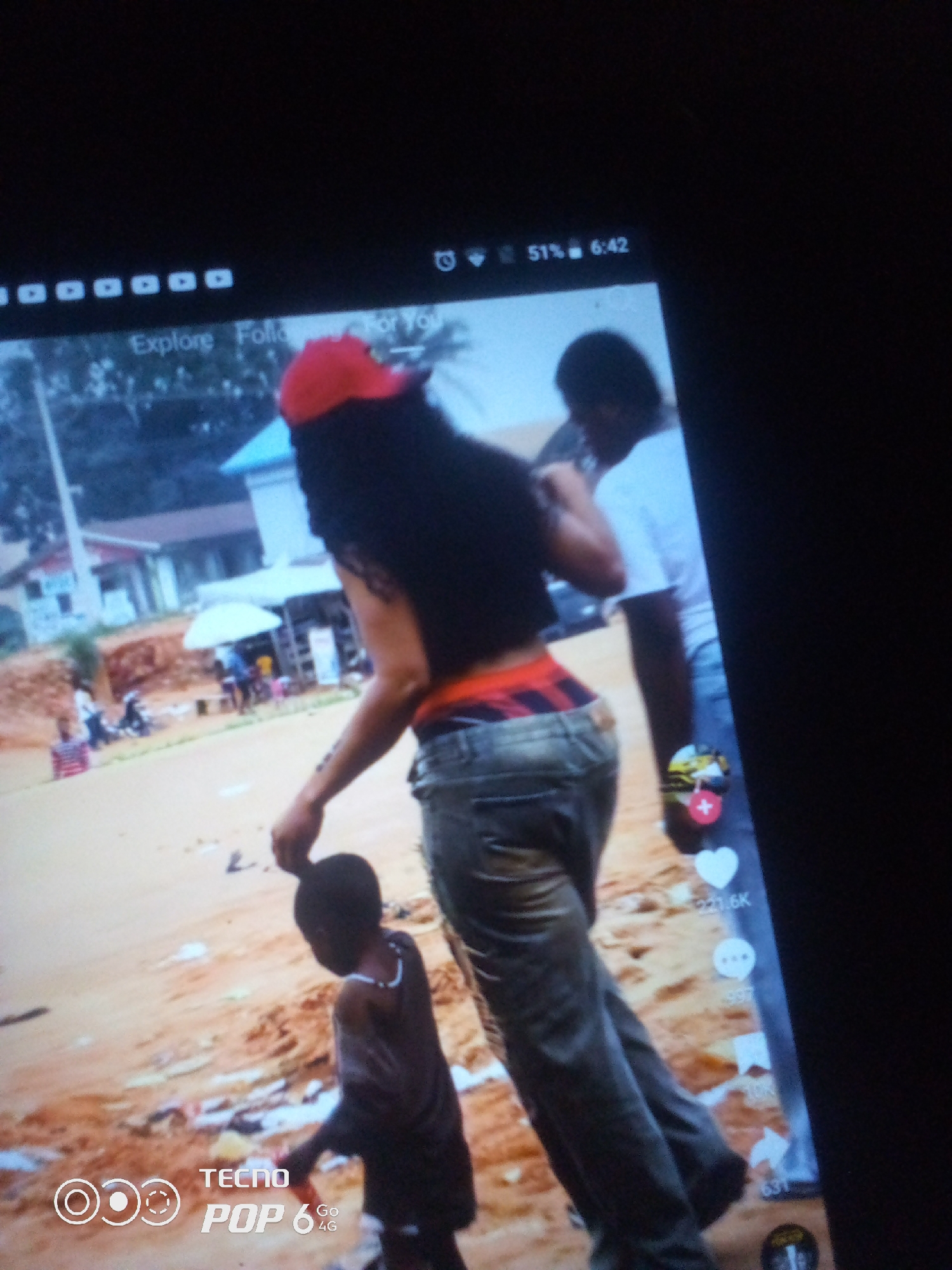 The image displays a screen showing a TikTok video. The video depicts two adults and a child in an outdoor environment. One adult, wearing a red cap, black long hair, and baggy pants, is facing away from the camera and appears to be interacting with the child. The other adult, in a white t-shirt, is standing behind the first adult. A young child, wearing a dark shirt, is in front of the adults, looking towards the ground. The background shows a sandy area with some buildings and a white umbrella, suggesting a semi-urban or rural setting. The screen also displays TikTok interface elements, including icons for likes, comments, shares, and the username "TECNO POP 6 Go 4G". The location is identified as Howayi, Nigeria.