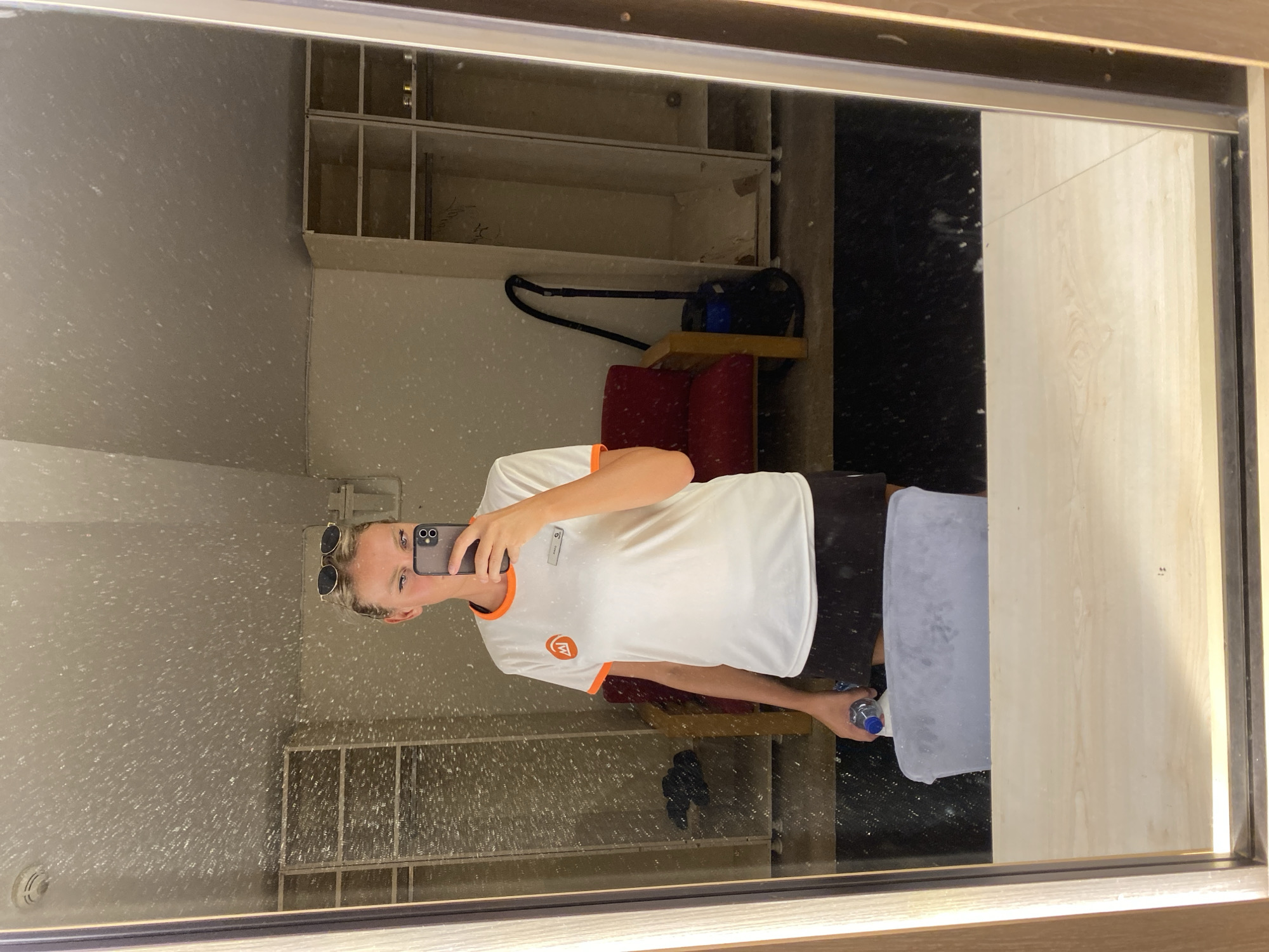 A woman with blonde hair pulled back and wearing sunglasses on her head is reflected in a mirror, taking a selfie with her phone. She is wearing a white t-shirt with orange trim and a black skirt. She is holding a water bottle in her left hand. The background appears to be a locker room or dressing room with open lockers, a red chair, and a vacuum cleaner. The mirror is dusty, with many speckles visible on its surface. The overall impression is of a backstage or service area. The location is not specified as Aksu, Turkey.