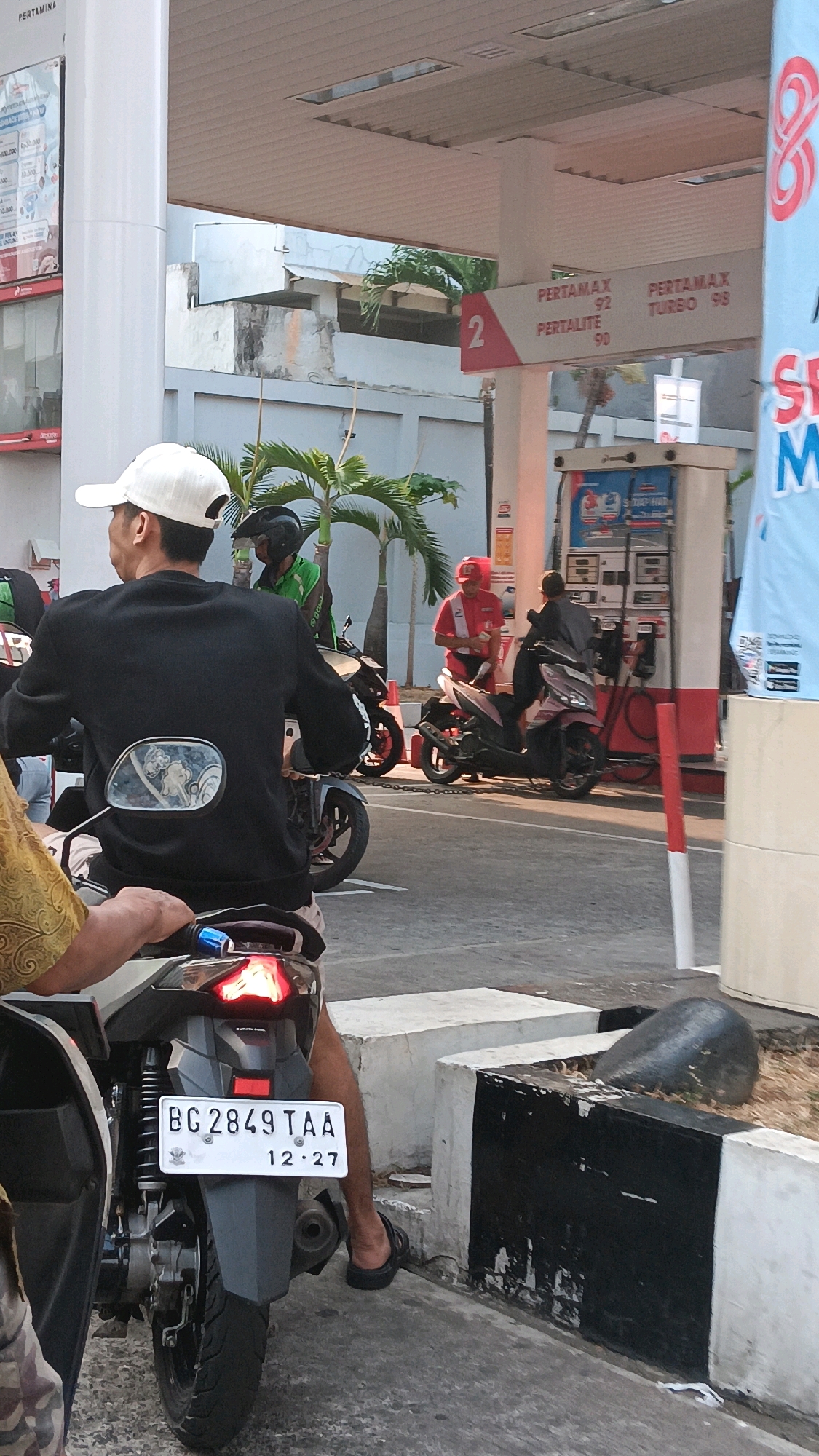 At a Pertamina gas station in South Jakarta, Indonesia, several individuals on motorcycles are visible. A man wearing a black jacket and a white cap is seated on a white motorcycle, with his back to the camera. Another person in a green uniform is on a motorcycle behind him, and a person in a red uniform, likely a gas station attendant, is seen near the gas pumps. The gas pumps are labeled with prices for "PERTAMAX 92", "PERTALITE 90", and "PERTAMAX TURBO 98". The scene appears to be a typical transaction at a gas station, with vehicles and personnel engaged in refueling activities. The ground is paved, and palm trees are visible in the background, suggesting a tropical climate. The license plate on the white motorcycle reads "BG 2849 TAA" and has the date "12.27" on it.