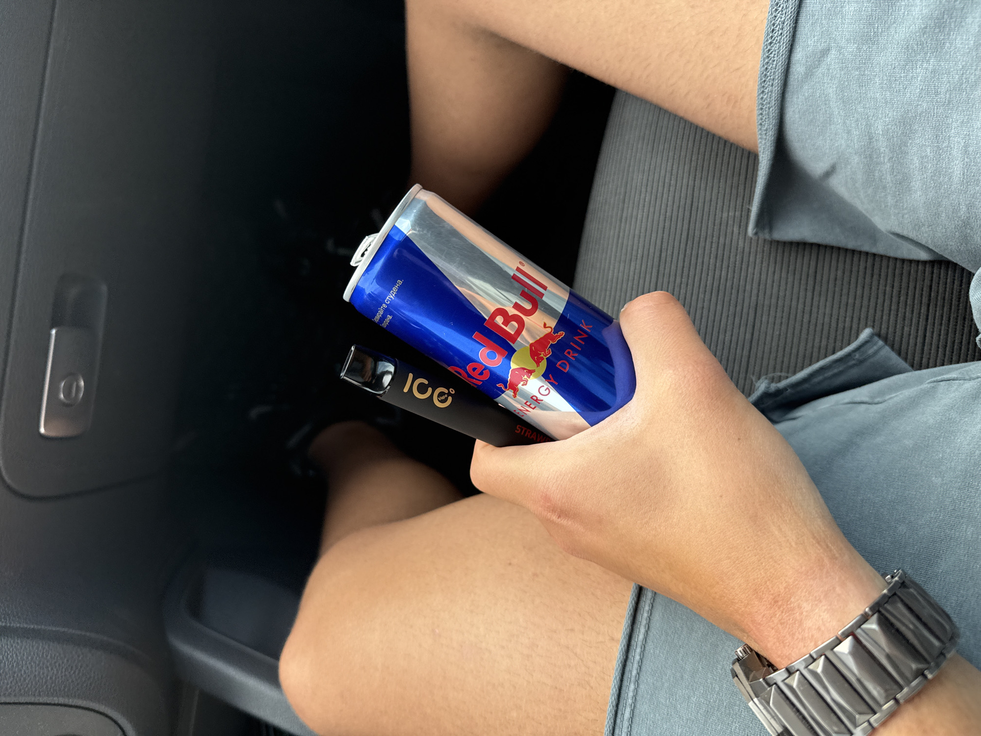 A person is holding a can of Red Bull and a black Icc Vape in what appears to be the interior of a car. The person is wearing a watch on their left wrist. The location is not evident from the image. No part of Hadzhidimitrovo, Bulgaria can be identified.