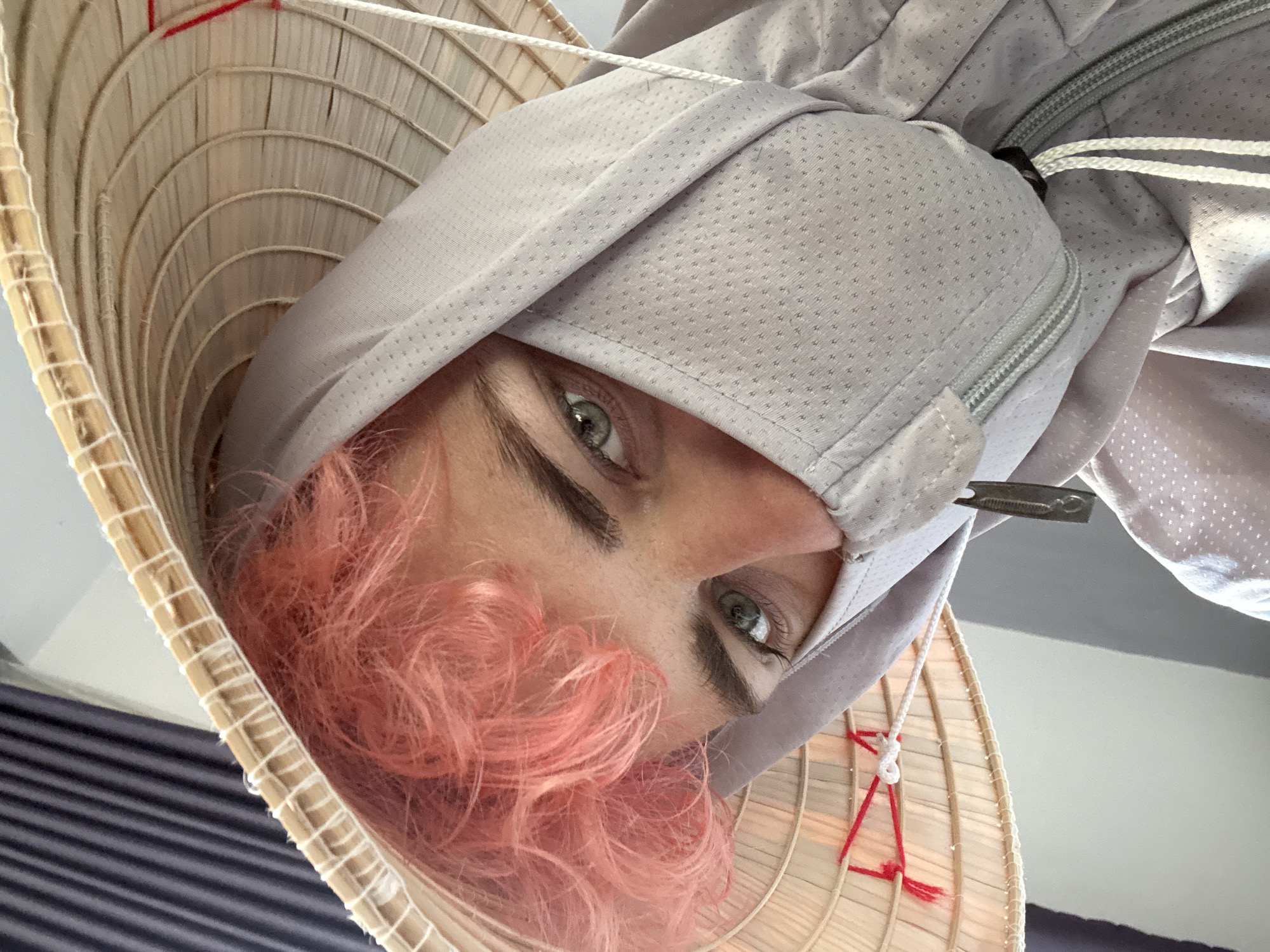 A person with pink hair and green eyes is wearing a traditional Vietnamese conical hat (nón lá) and a light gray hooded jacket. The person is looking directly at the camera. The scene appears to be indoors. The background is blurred. There is no definitive information in the image to identify Đà Nẵng as the location.