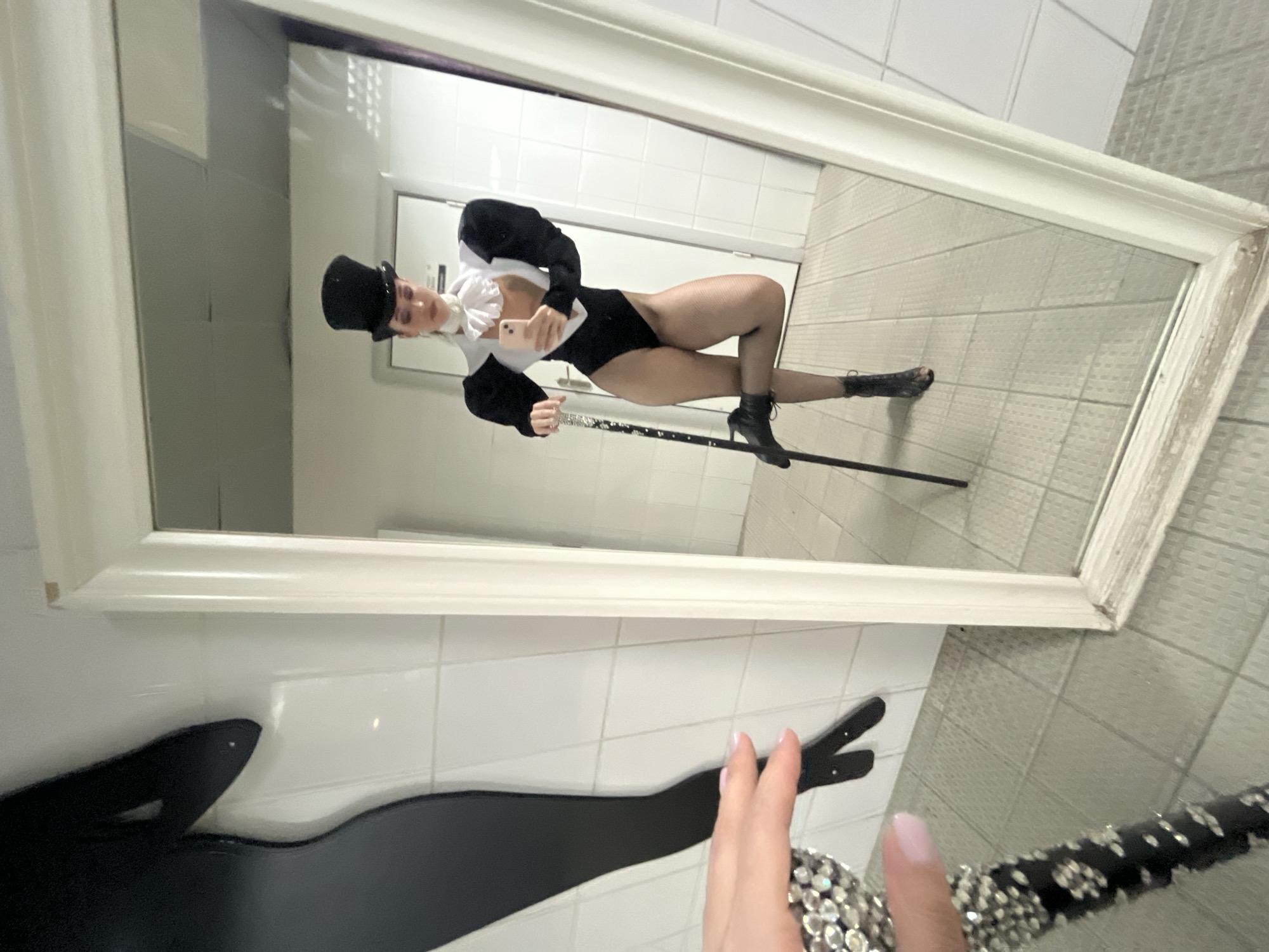 A person is posing for a selfie in a mirror while holding a bejeweled cane. The person is wearing a black and white theatrical costume that includes a leotard, puffed sleeves, a ruffled collar, fishnet stockings, and a top hat. They are also wearing black ankle boots and standing with one leg bent. The reflection shows the person in what appears to be a backstage area with white tiled walls and a tiled floor. The environment does not provide any specific clues to identify the location as Kazan, Russia.