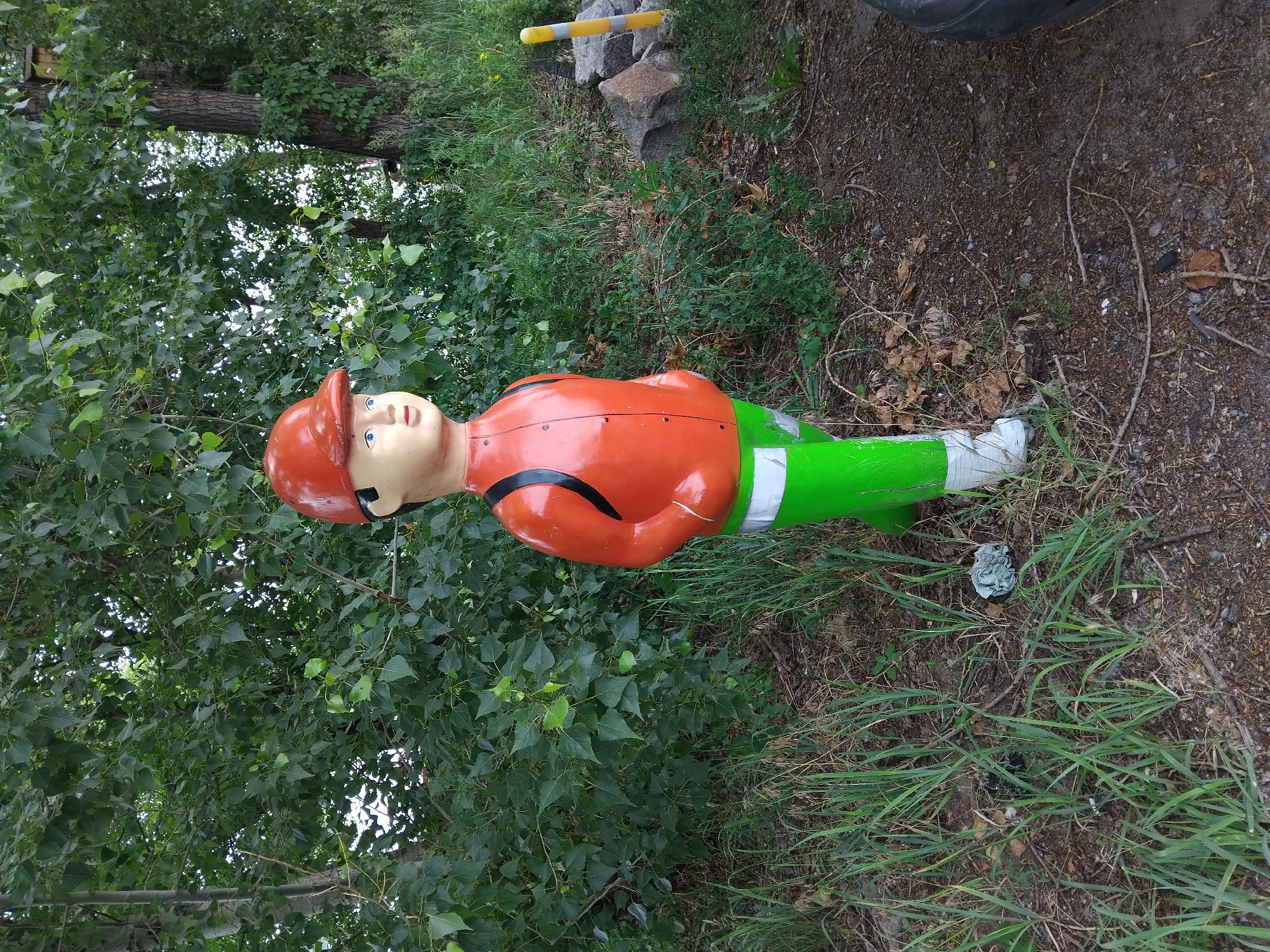 An outdoor, eye-level, full-shot photograph shows a full-body statue of a child wearing a red cap, orange jacket, and green pants. The statue is positioned in the center of the frame, standing on a patch of dirt and grass. The statue is facing right, with its hands clasped behind its back. A black strap is visible across the statue's chest. The statue's pants have a white reflective band around the lower calf, and its feet are clad in white boots. The background consists of lush green foliage, with trees and bushes extending upwards and to the left of the statue. To the right of the statue, there is a yellow and gray striped post, some rocks, and dense green vegetation. The ground in front of the statue is covered in grass and dirt, with some fallen leaves scattered around. A portion of a black tire is visible in the lower right corner of the image. The scene suggests an outdoor location, possibly a park or a roadside, in a natural setting.