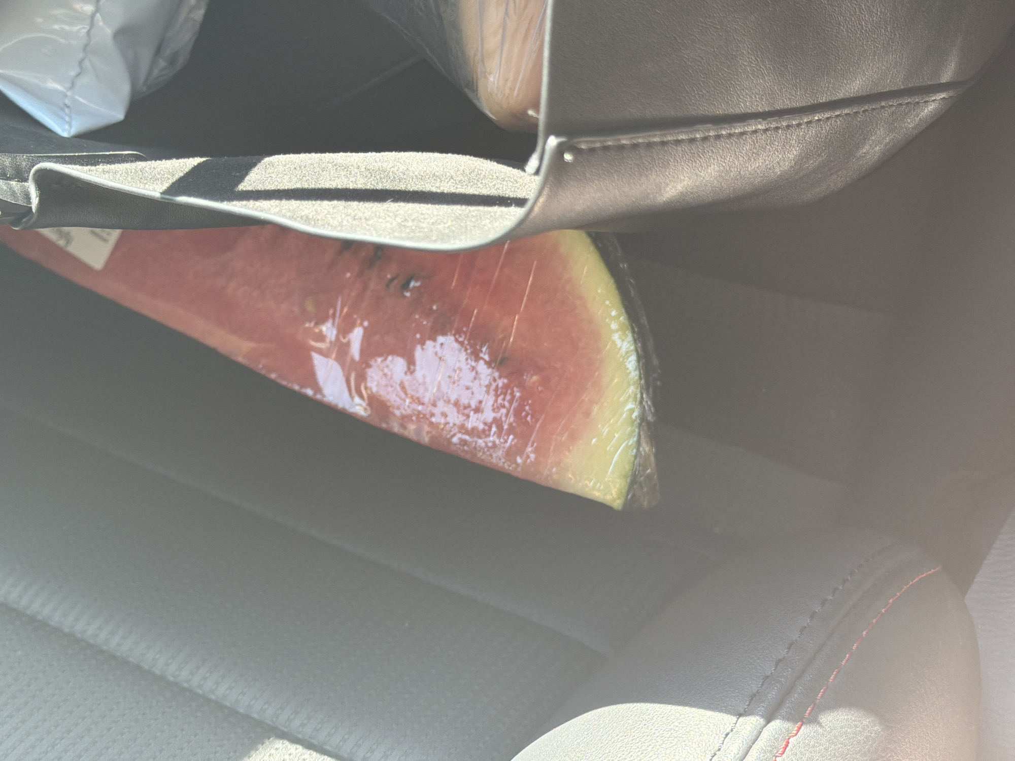 A wedge of watermelon, wrapped in plastic, sits on a car seat. A black leather bag sits above and slightly obscures the watermelon. The car seat is black leather with red stitching.