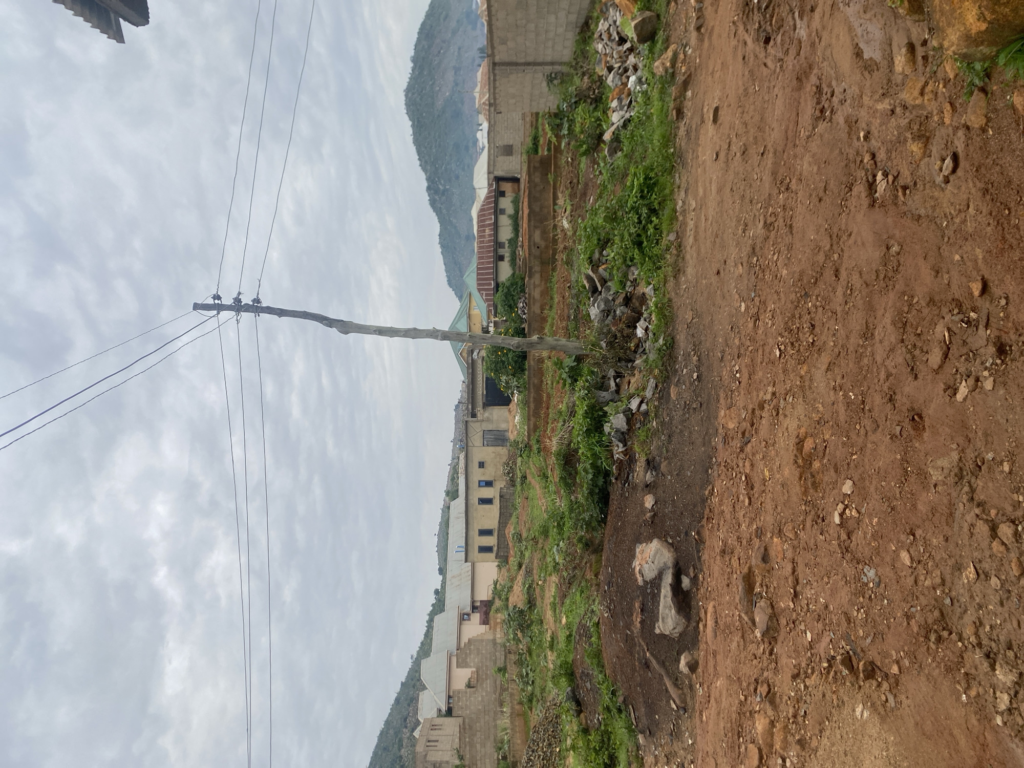 In the city of Jos, Nigeria, an overcast sky looms above a developing area featuring several unfinished and completed buildings. A wooden utility pole stands prominently in the foreground, with electrical wires stretching across the frame. The terrain is uneven and dusty, with piles of rocks and construction debris scattered throughout the scene. Green vegetation sprouts in patches, indicating ongoing growth amidst the construction. The backdrop is dominated by rolling hills and mountains, characteristic of Jos's natural landscape. The construction activity suggests a neighborhood in the process of expansion or renovation, with a mix of residential structures in various stages of completion.