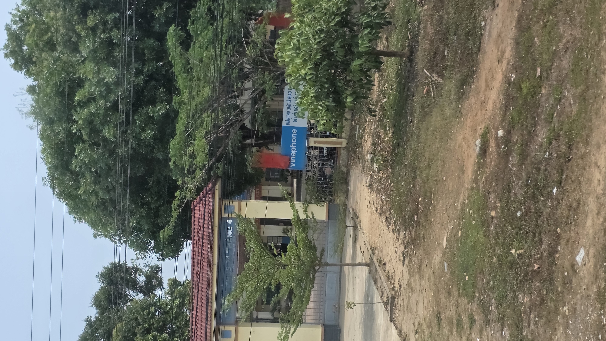 The image shows a slightly angled view of a building located in Tân Cảnh, Vietnam. The building appears to be a school or community center, with a red-tiled roof and yellow walls. A sign for "vinaphone" is visible near the entrance. The surrounding area consists of trees, grass, and a dirt path leading to the building. Overhead, power lines crisscross the sky. There are no people visible in the image.