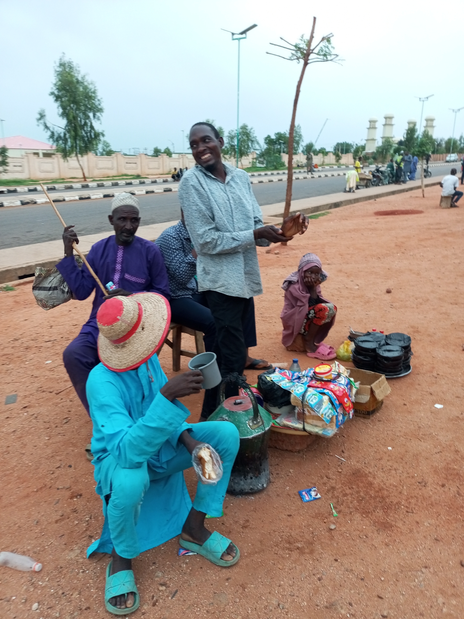 In Gombe, Nigeria, a group of individuals are gathered near a dusty roadside. In the foreground, a person in a bright blue traditional outfit, adorned with a straw hat with a red band, sits and holds a cup. Another person, dressed in purple, sits with a walking stick and a bag. A third individual, wearing a light blue shirt, stands and appears to be interacting with the seated group. To the right, a young child in a pink headscarf and dress sits on the ground, looking thoughtful. Various items are scattered around, including a stack of black bowls, plastic containers, bags of goods, and a discarded plastic bottle. In the background, a street with a sidewalk and sparse trees is visible, with a building and some white, domed structures in the distance. The sky is overcast, indicating a cloudy day. The overall scene depicts a casual roadside gathering in a Nigerian urban setting.