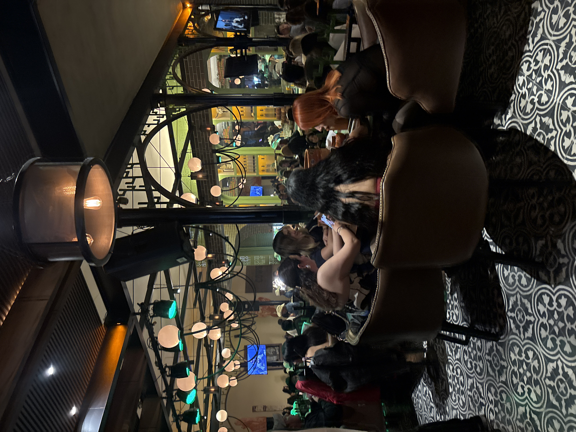 The image captures a bustling interior scene, likely in a bar or restaurant. The location is implied to be in Puebla, Mexico.
Several people are seated and engaged in conversations, possibly dining, while some are using their phones. The lighting is dim and atmospheric, with decorative globe lights and industrial-style lamps hanging from the ceiling. Arched architectural elements are visible in the background.
The foreground features brown leather chairs and a floor with a black and white patterned tile. There are TVs visible in the background with screens turned on.
