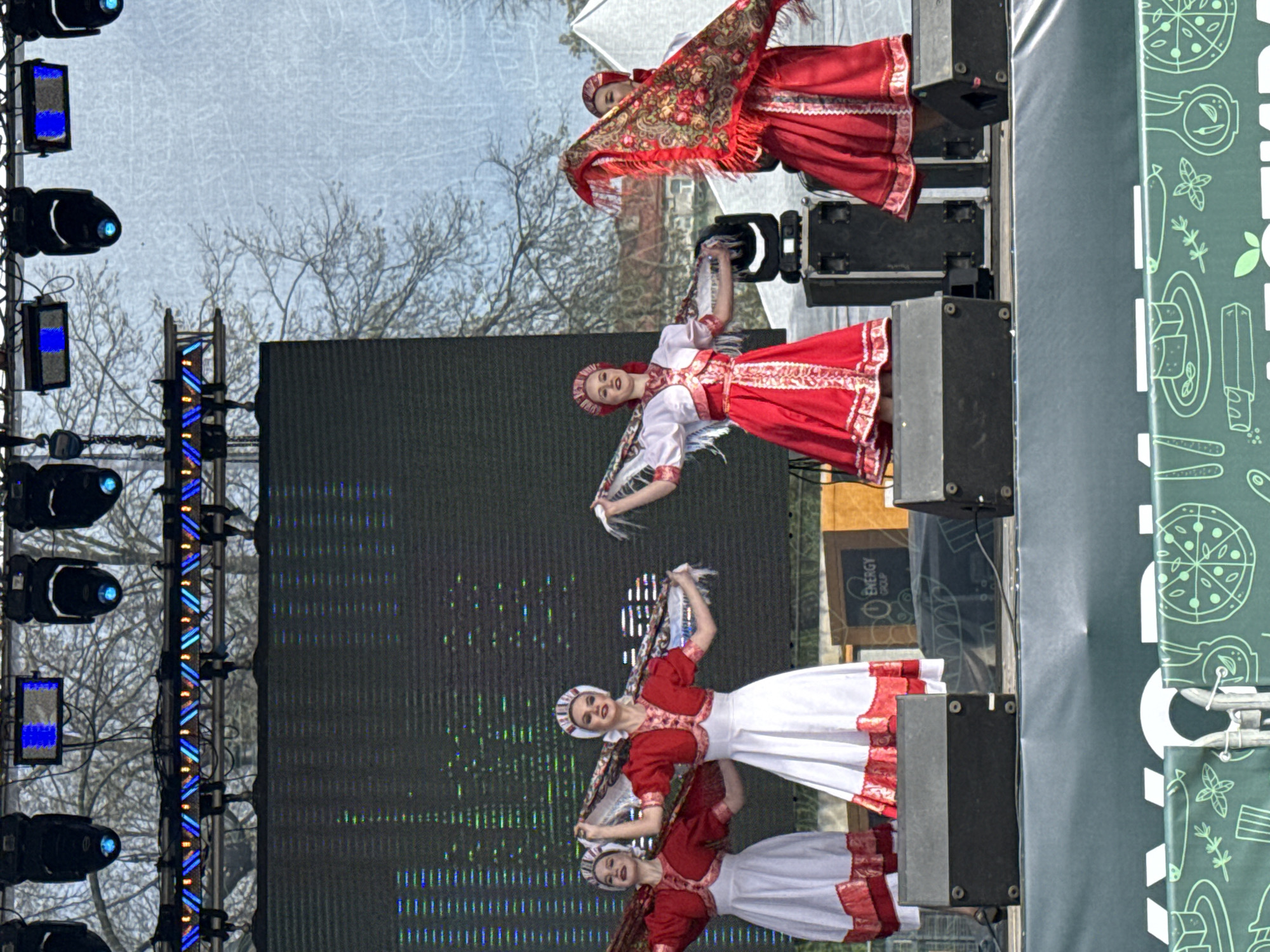 On an outdoor stage, a group of women dressed in traditional Russian folk costumes performs a dance. The women are in red and white dresses with headscarves. They hold scarves as they perform. To the left of the stage, there is lighting equipment. In the background, trees are visible. To the right of the stage, there is signage. The location is suggested to be Saint Petersburg, Russia.