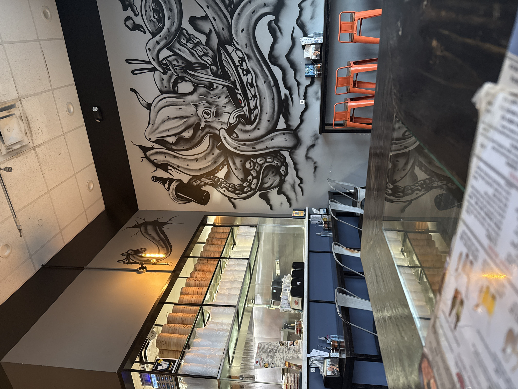 Here's a detailed description of the image:
**Location:** The image appears to be taken inside a restaurant or bar in Dania Beach, United States of America.
**Environment:**
* The interior features a modern design.
* There is a prominent mural on the wall depicting a large octopus.
* There are metal frame shelves storing cups and supplies, plus, behind the cups, there are napkins.
* The space appears well-lit.
**Objects:**
* Mural of an octopus with prominent tentacles.
* Rows of orange barstools are fixed to the wall.
* Metal frame shelves containing cups, napkins, and other supplies.
* There are tall silver chairs along the bar.
* Behind the cups and napkins, there's a glass enclosure that is part of the shelves.
* There is a cash register with some cards and a payment terminal on the counter next to the shelves.
**Subjects:**
* The primary subject is the interior design and layout of the establishment.
**Actions and Interactions:**
* The scene is static, showing the establishment's interior.
**Overall:** The image provides a view of a unique and stylized interior, showcasing a combination of functional storage and artistic elements.