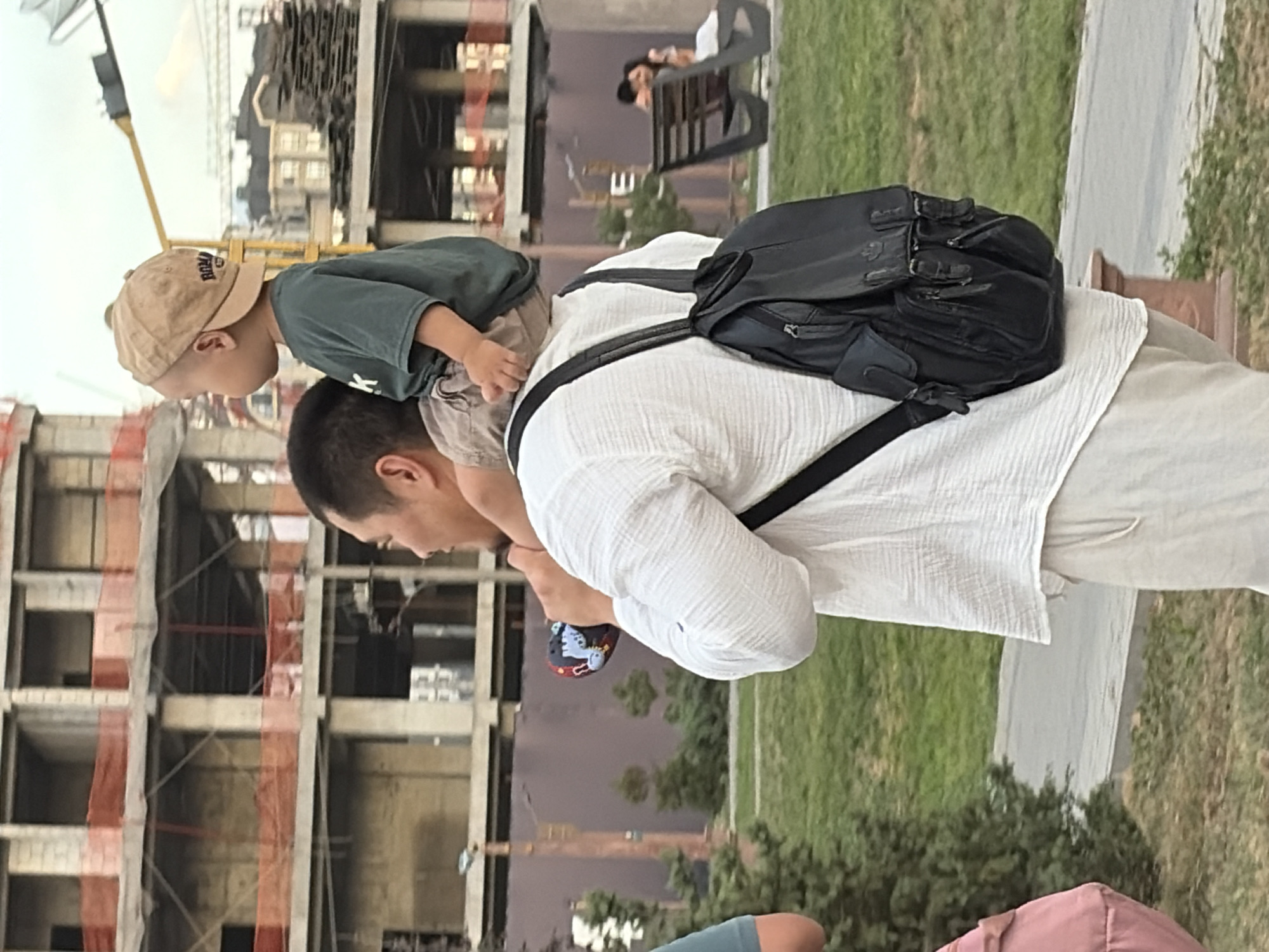 An adult male wearing a white linen shirt and light-colored pants, carrying a black backpack, walks with a young child on his shoulders. The child wears a green t-shirt and a tan baseball cap. In the background, construction is underway on a multi-story building with exposed concrete and scaffolding. To the right, a park bench is occupied by a couple. The environment appears to be an urban park setting.