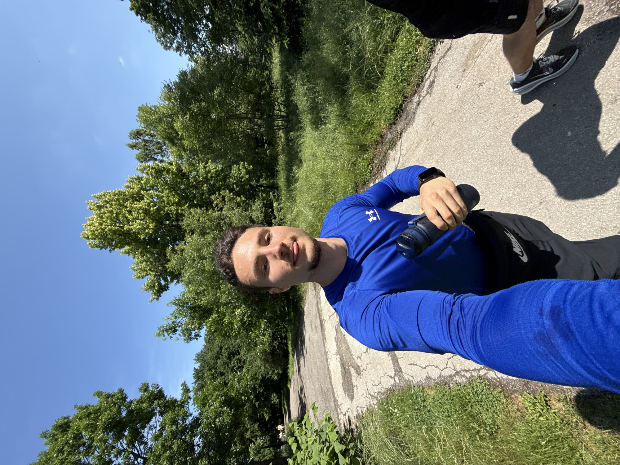 This rotated outdoor selfie captures a man on a sunny day in what appears to be a park or green space in Sofia, Bulgaria. He is wearing a blue long-sleeved athletic shirt with an "Under Armour" logo on the chest, black shorts, and a black smartwatch on his left wrist. He holds a dark water bottle in his left hand and looks towards the camera with a neutral expression. Some perspiration is visible on his blue sleeve, suggesting physical activity.
The setting includes a cracked asphalt or concrete path, bordered by lush green grass, various bushes, and numerous trees with vibrant green foliage under a clear, bright blue sky with a few wispy clouds. The strong sunlight casts distinct shadows on the path, indicating a mid-morning or afternoon time.
Partially visible in the upper right corner are the legs and feet of another person, wearing black shorts and dark athletic shoes with a "Nike" logo, suggesting they are accompanying the man on a walk or run. The overall scene conveys an active outdoor moment in a natural, well-lit environment.