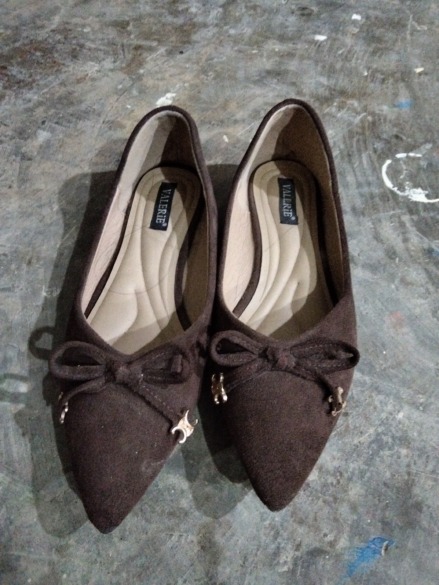 A pair of brown, pointed-toe ballet flats with a bow detail on the toe is displayed on a textured, grey surface that appears to be concrete. The shoes have a light brown insole with a black tag bearing the white text "VALERIE". Small gold-colored charms are attached to the sides of the bow on each shoe. The surface shows some white and blue marks, suggesting an outdoor or workshop environment. The image is taken from a top-down perspective.