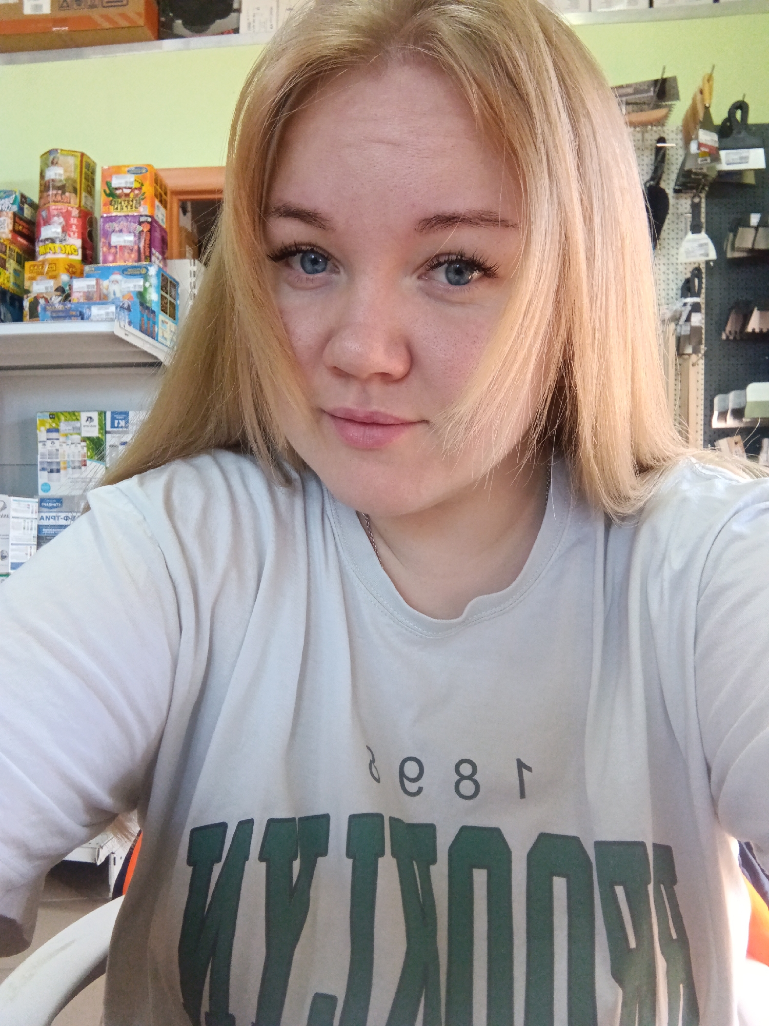 A young woman with fair skin, blue eyes, and blonde hair stands in a well-lit interior. Some of her hair falls across her face. She is wearing a white t-shirt with the word "BROOKLYN" printed in dark green. The letters are slightly distorted, appearing backward. The location appears to be a store, with shelves stocked with various products visible in the background, including what appears to be fireworks and other merchandise. The background also includes a green wall with hanging tools or hardware. The location of the store is Фоки, Russia.