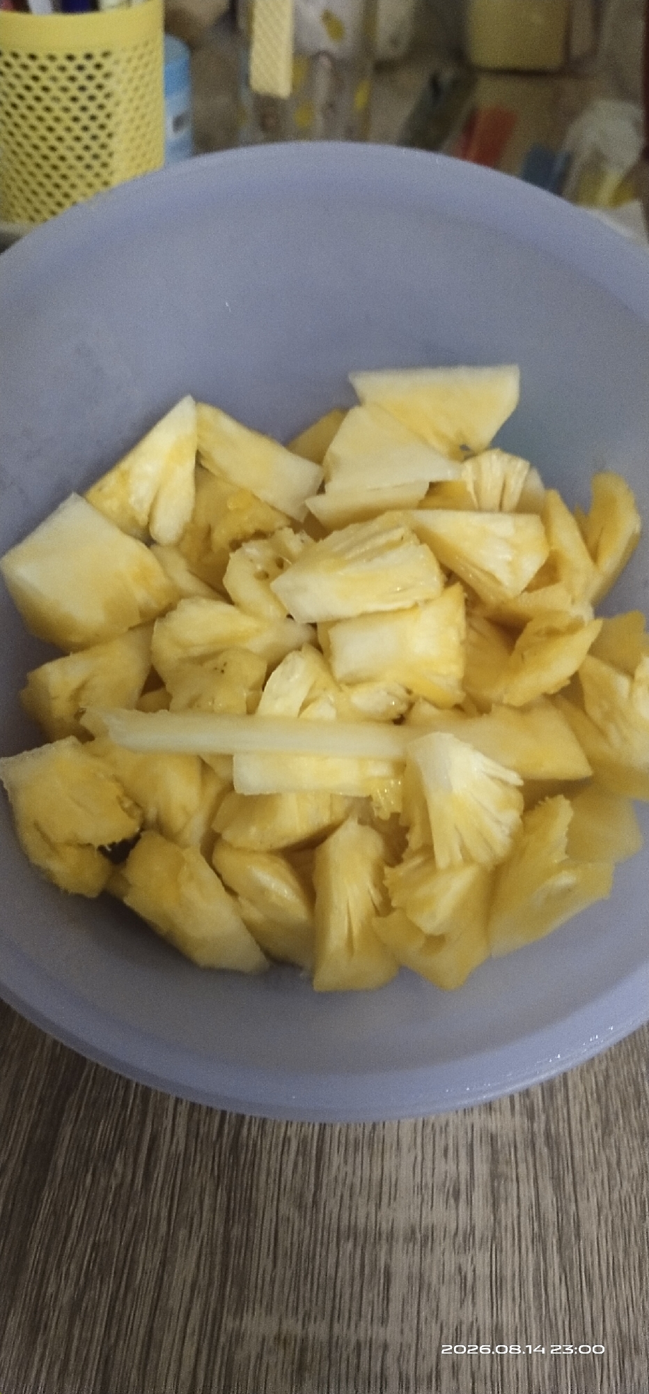 A bowl of sliced pineapple is placed on a wooden surface. The bowl is light purple and filled with numerous pieces of ripe pineapple, cut into bite-sized chunks. The pineapple is a vibrant yellow with some lighter yellow flesh visible. The wooden surface has a visible grain pattern. In the background, out of focus, are some yellow and blue items on a shelf. The scene appears to be indoors, likely in a kitchen or dining area. The timestamp "2026.08.14 23:00" is visible in the bottom right corner. There are no people or animals present in the image.